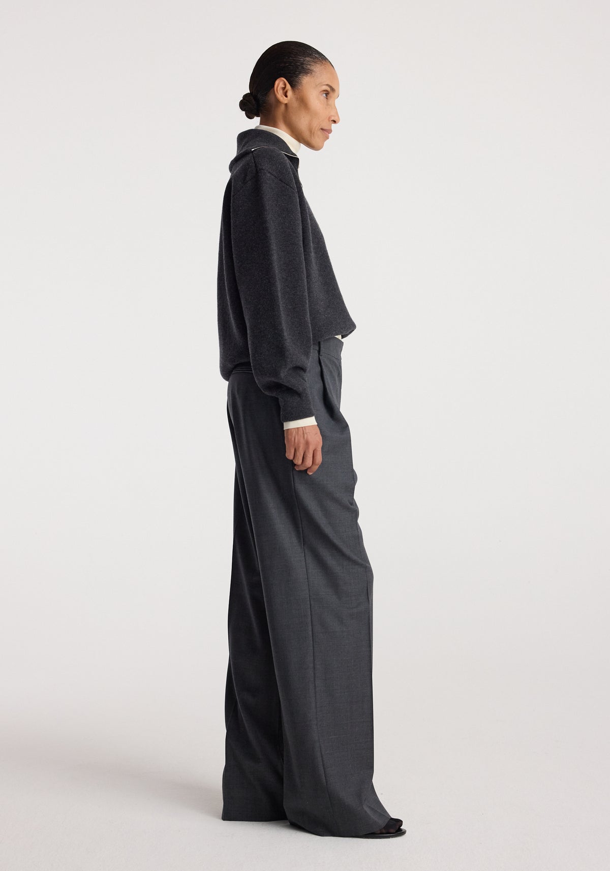 Woman wearing a charcoal color Two-way zipped turtleneck | charcoal with long sleeves and a high neck, paired with dark grey wide-leg trousers, standing in profile against a plain background.