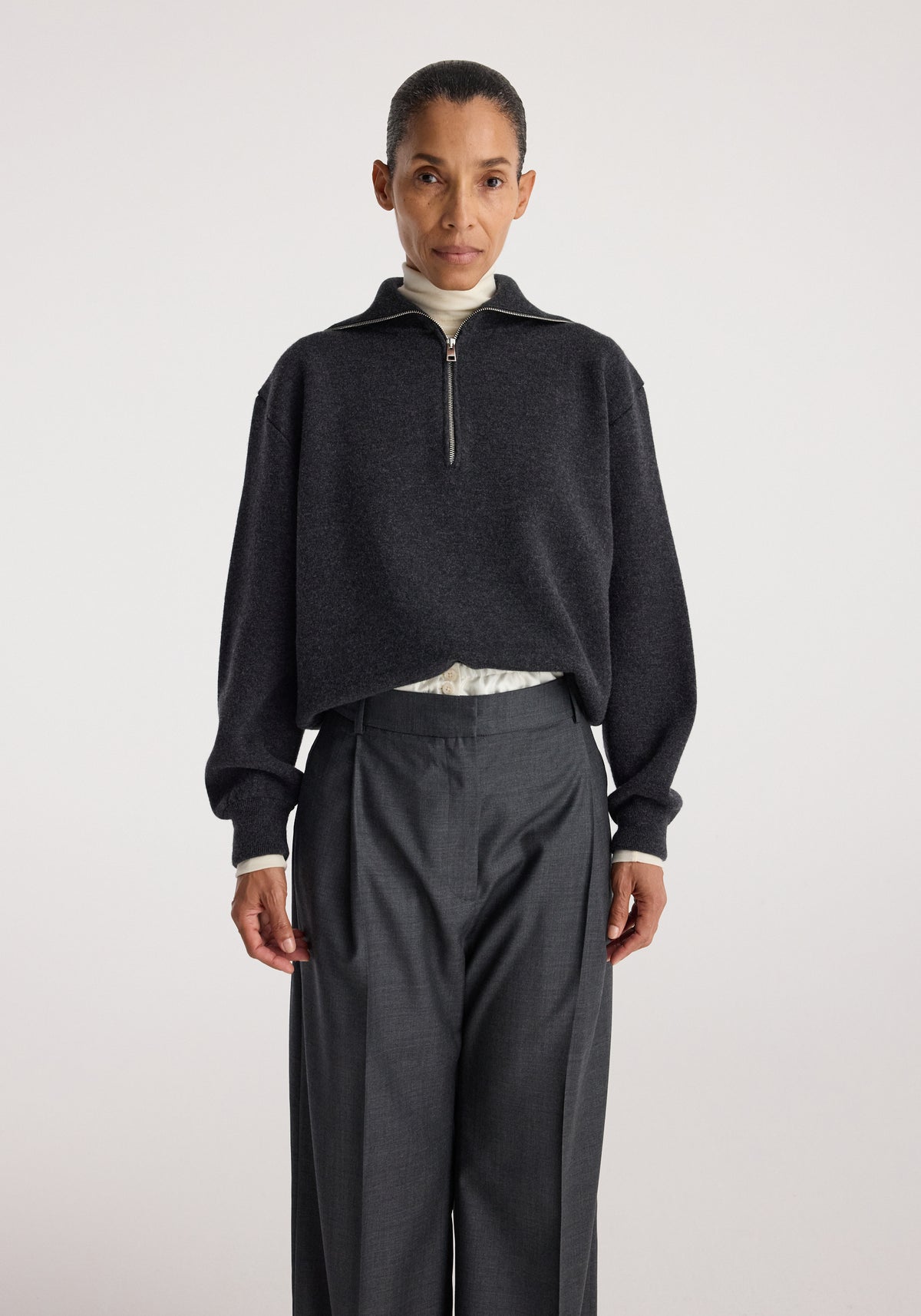 Woman wearing a charcoal Two-way zipped turtleneck with long sleeves and a loose fit, paired with high-waisted dark trousers against a plain background.
