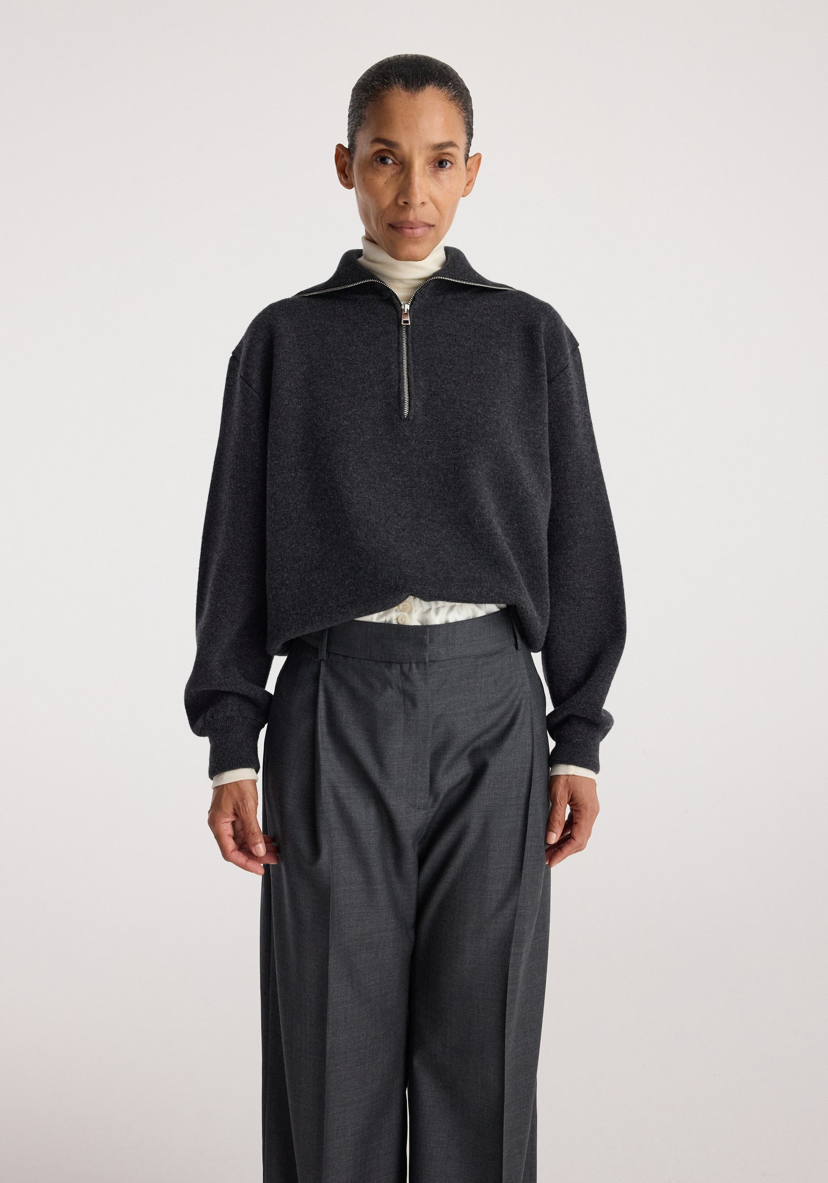 Woman wearing a charcoal Two-way zipped turtleneck with long sleeves and a loose fit, paired with high-waisted dark trousers against a plain background.