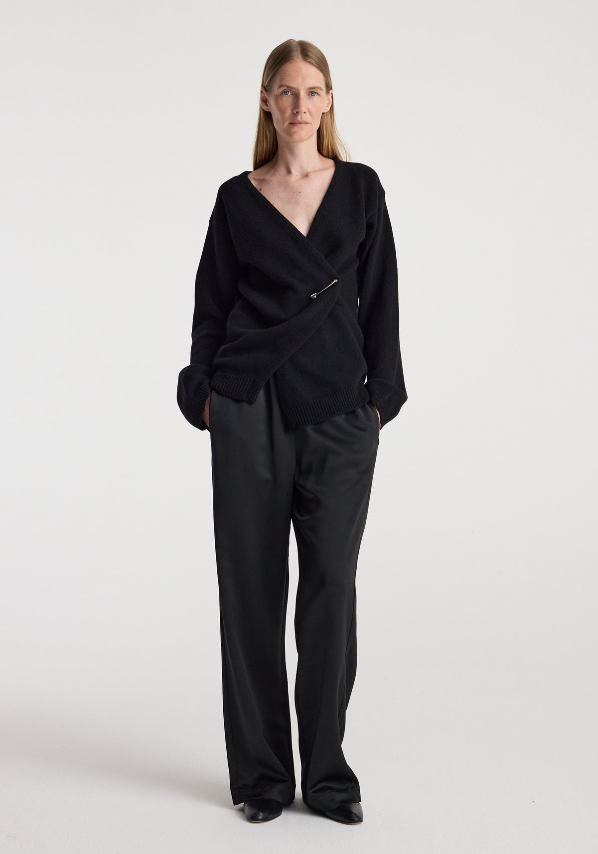 Woman wearing an elegant black off-shoulder wrap cardigan | black with relaxed black pants, standing against a plain white background.