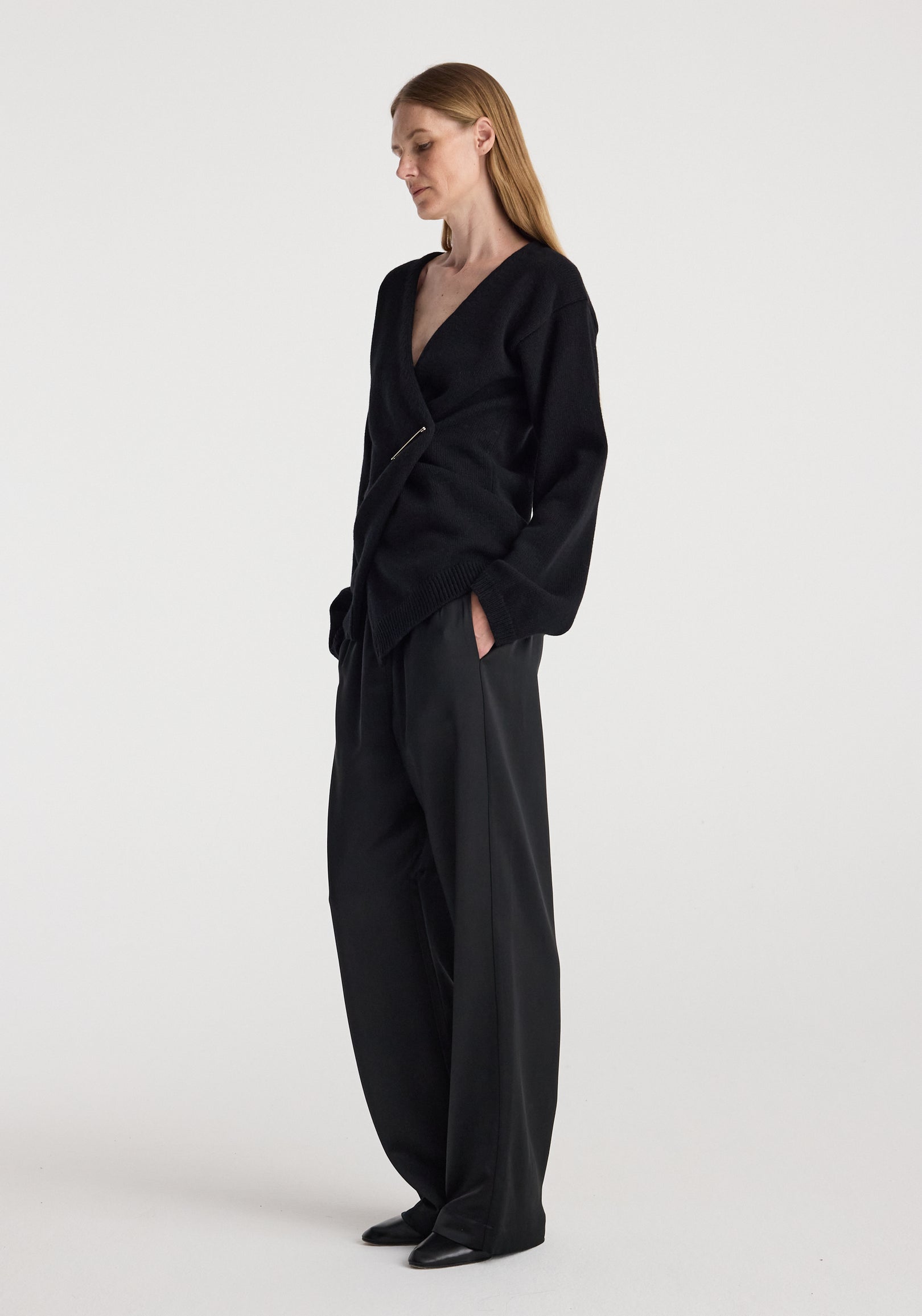 Woman wearing an off-shoulder wrap cardigan | black styled with loose black pants and black shoes, standing against a plain white background.