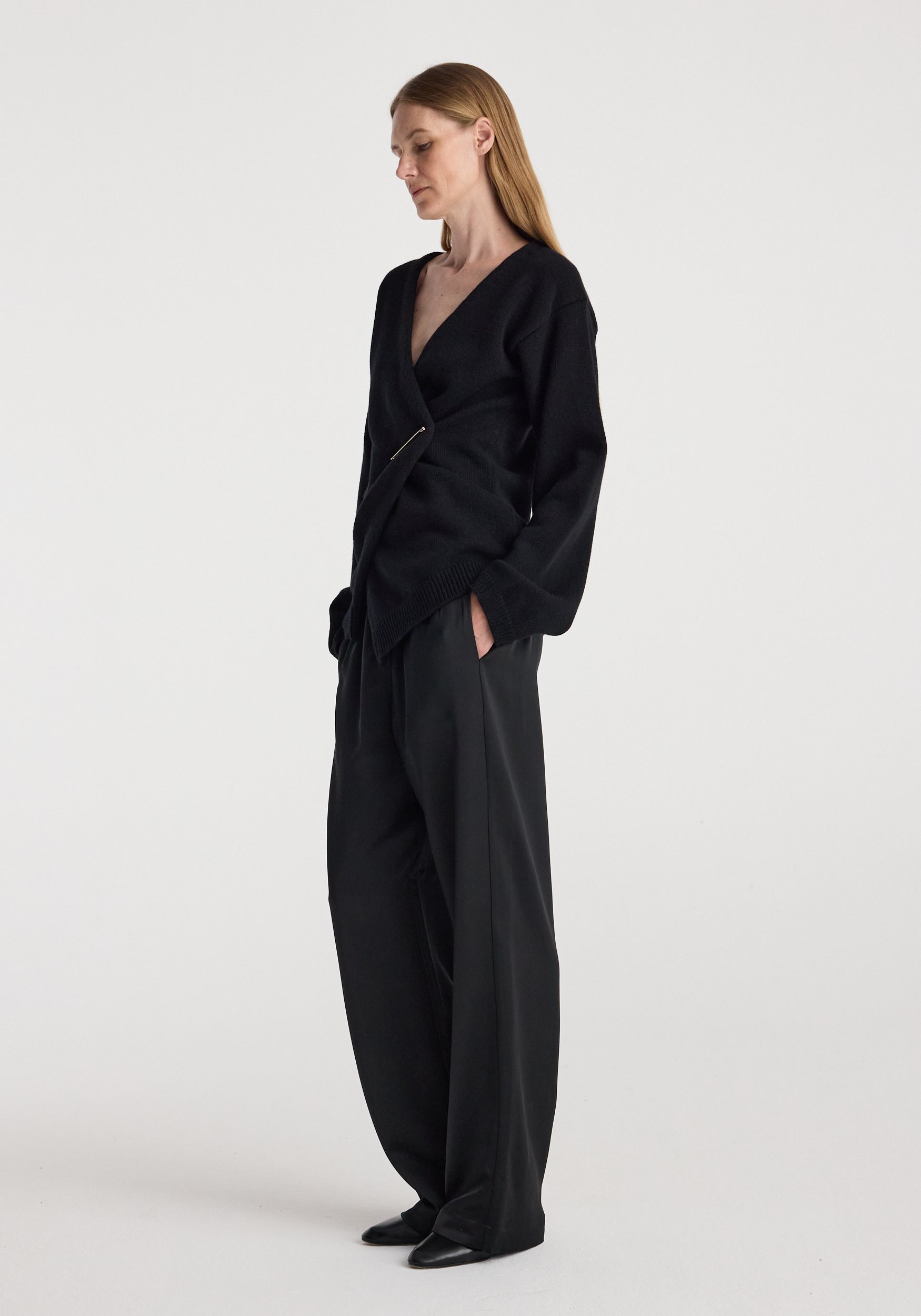Woman wearing an off-shoulder wrap cardigan | black styled with loose black pants and black shoes, standing against a plain white background.