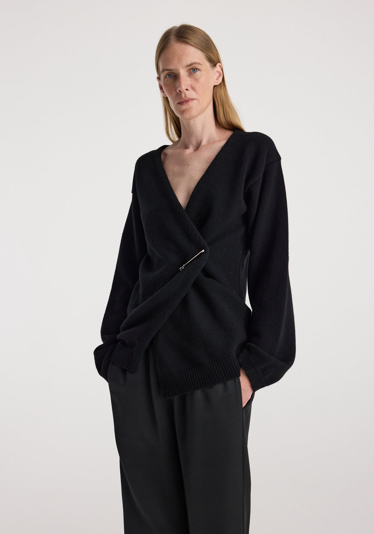 Woman wearing an off-shoulder wrap cardigan | black with a deep V neckline and long sleeves, styled with loose black pants against a plain background.