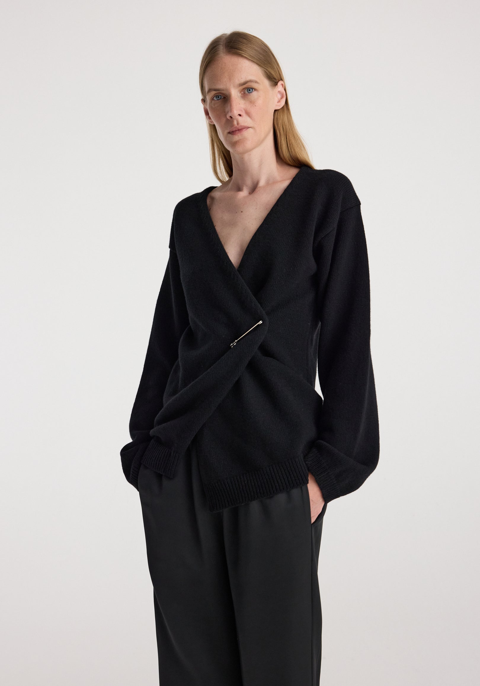 Woman wearing an off-shoulder wrap cardigan | black with a deep V neckline and long sleeves, styled with loose black pants against a plain background.