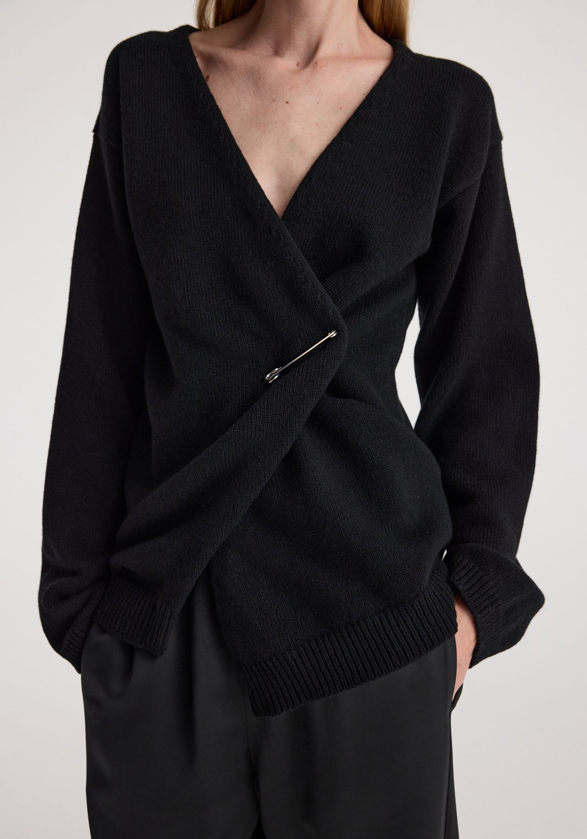 Woman wearing an Off-shoulder wrap cardigan | black with a deep V-neck and long sleeves, fastened with a safety pin detail in the front.
