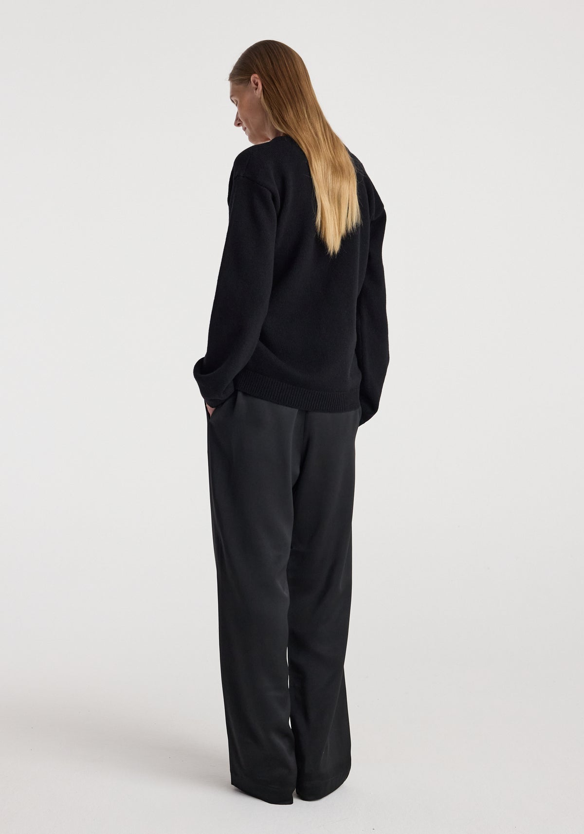 Woman standing with hands in pockets wearing black wide-leg pants and an off-shoulder wrap cardigan | black, viewed from the back against a plain background.