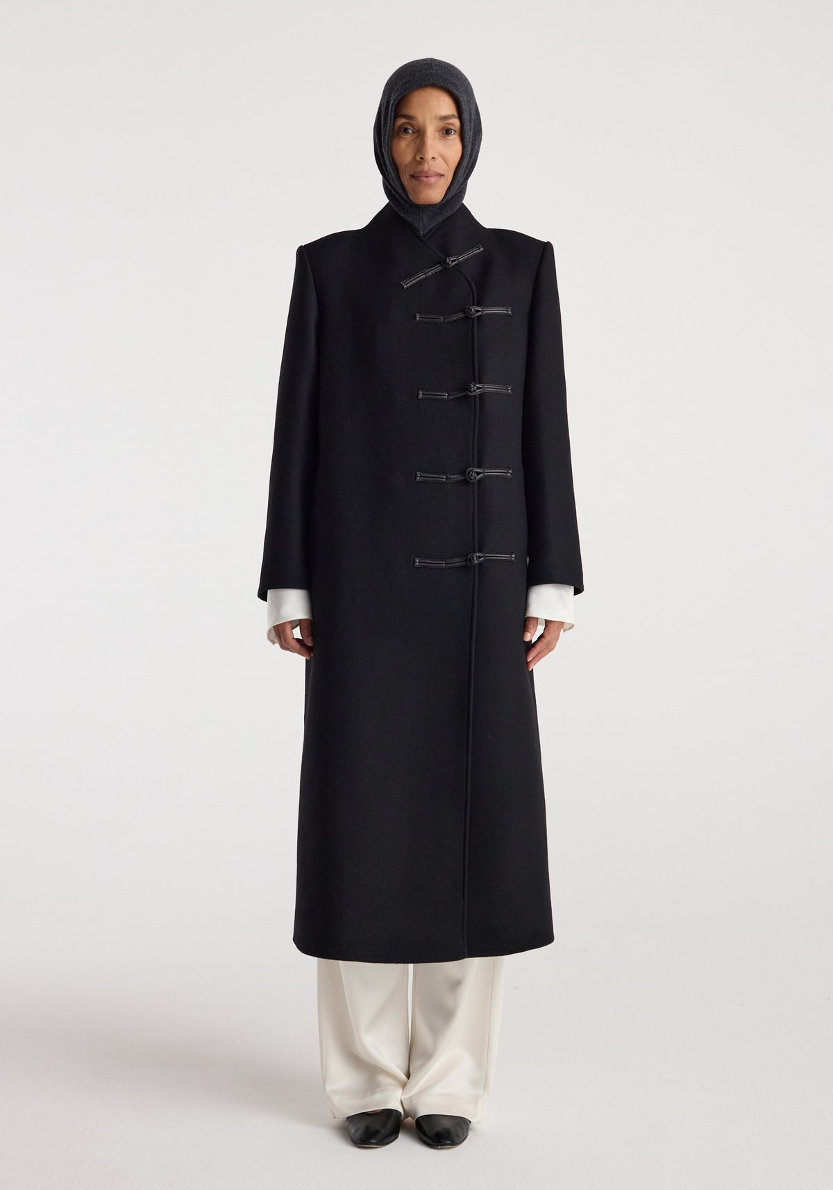 Woman wearing a black long coat with toggle closures and a Balaclava | anthracite melange head covering against a plain light background.