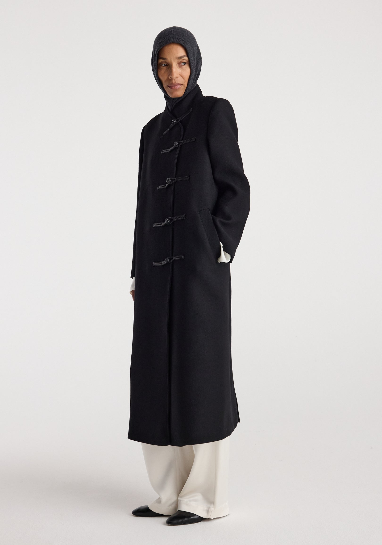 Woman wearing a dark coat and a Balaclava | anthracite melange hood covering her head and neck, standing against a plain light background.