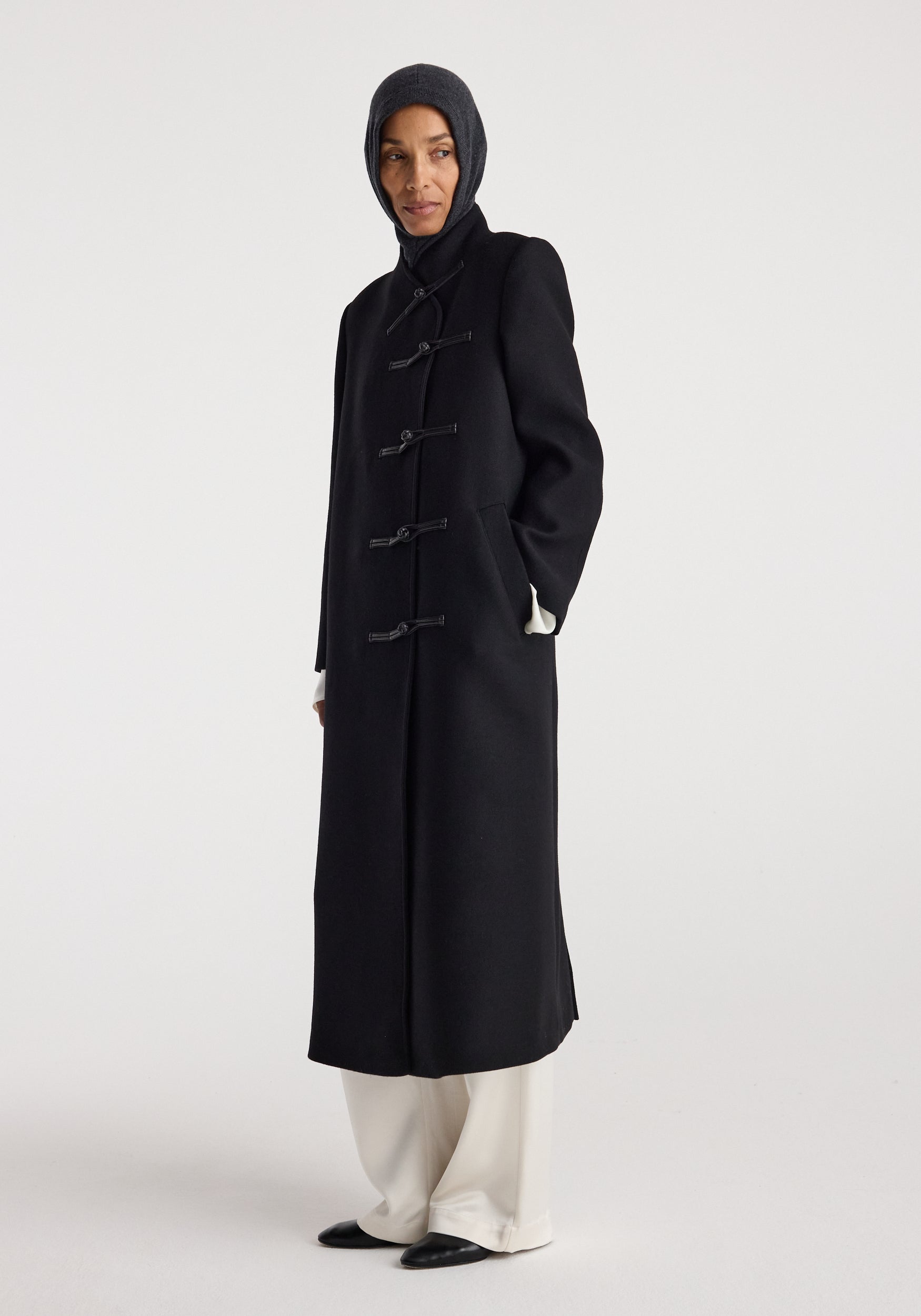 Woman wearing a dark coat and a Balaclava | anthracite melange hood covering her head and neck, standing against a plain light background.