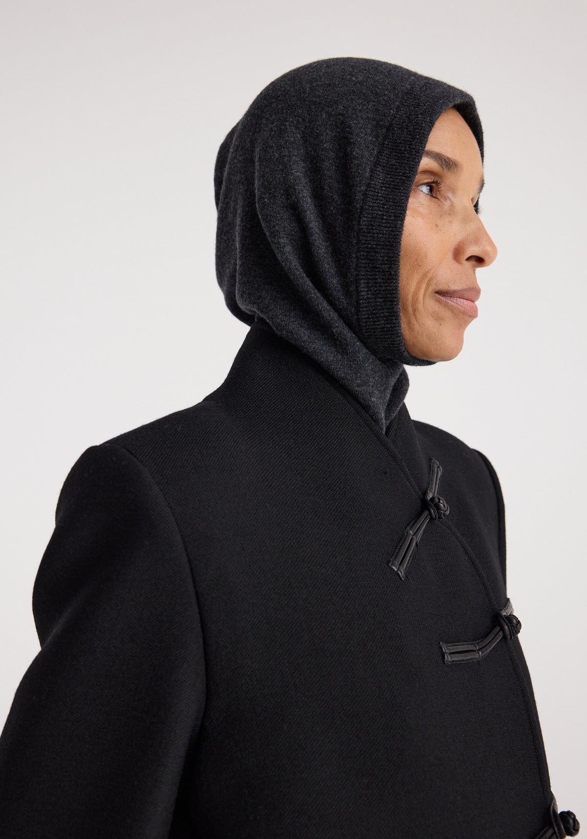 Profile of a person wearing a dark smooth-textured hood with a fitted design in Balaclava | anthracite melange, paired with a black coat featuring toggle closures.