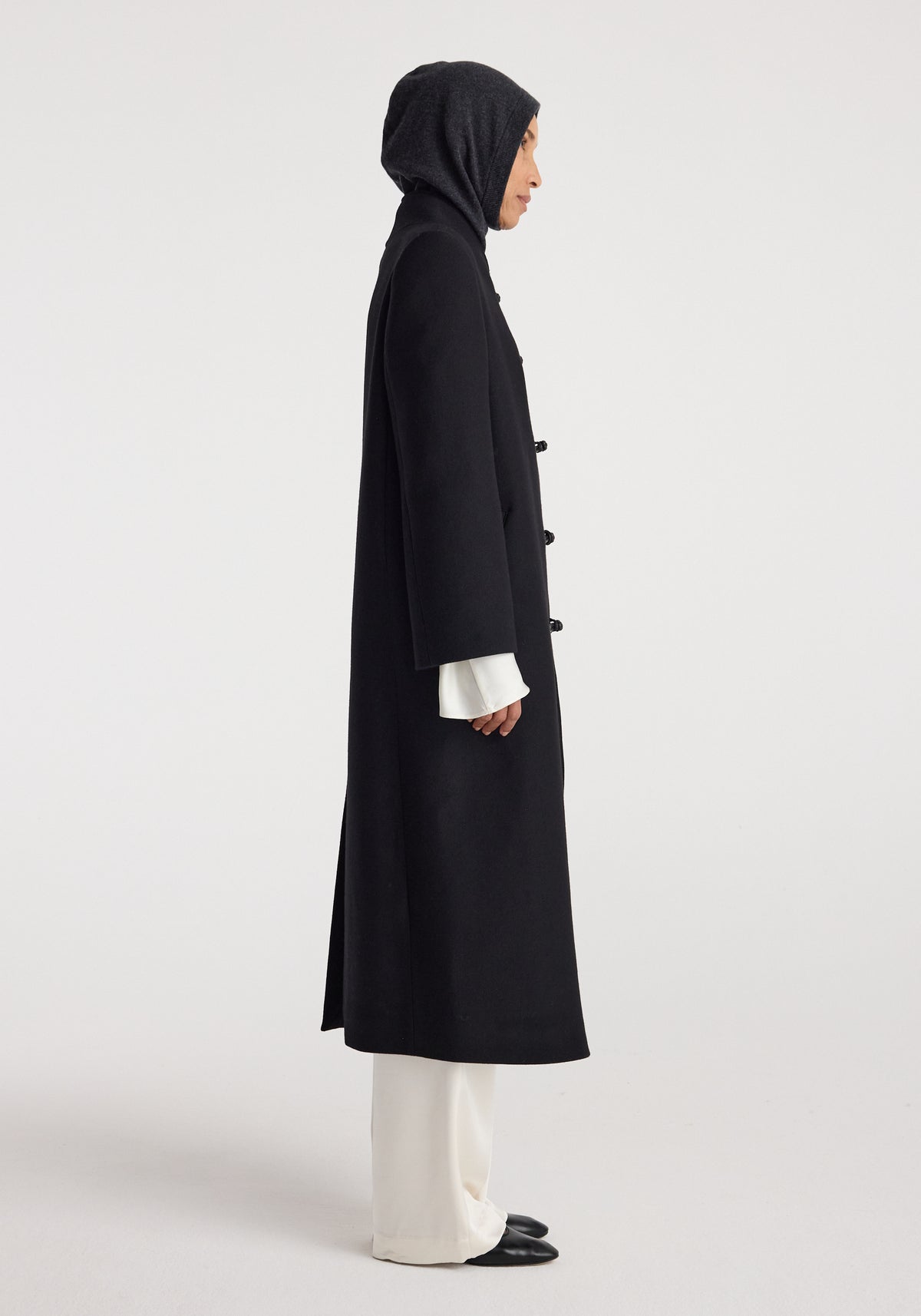 Side view of a person wearing a dark gray knit balaclava | anthracite melange paired with a long black coat, white wide-leg trousers, and black shoes against a plain background.
