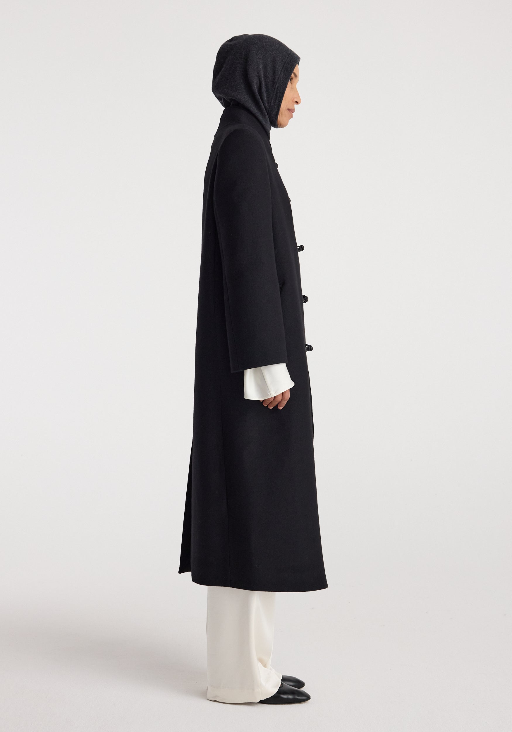 Side view of a person wearing a dark gray knit balaclava | anthracite melange paired with a long black coat, white wide-leg trousers, and black shoes against a plain background.