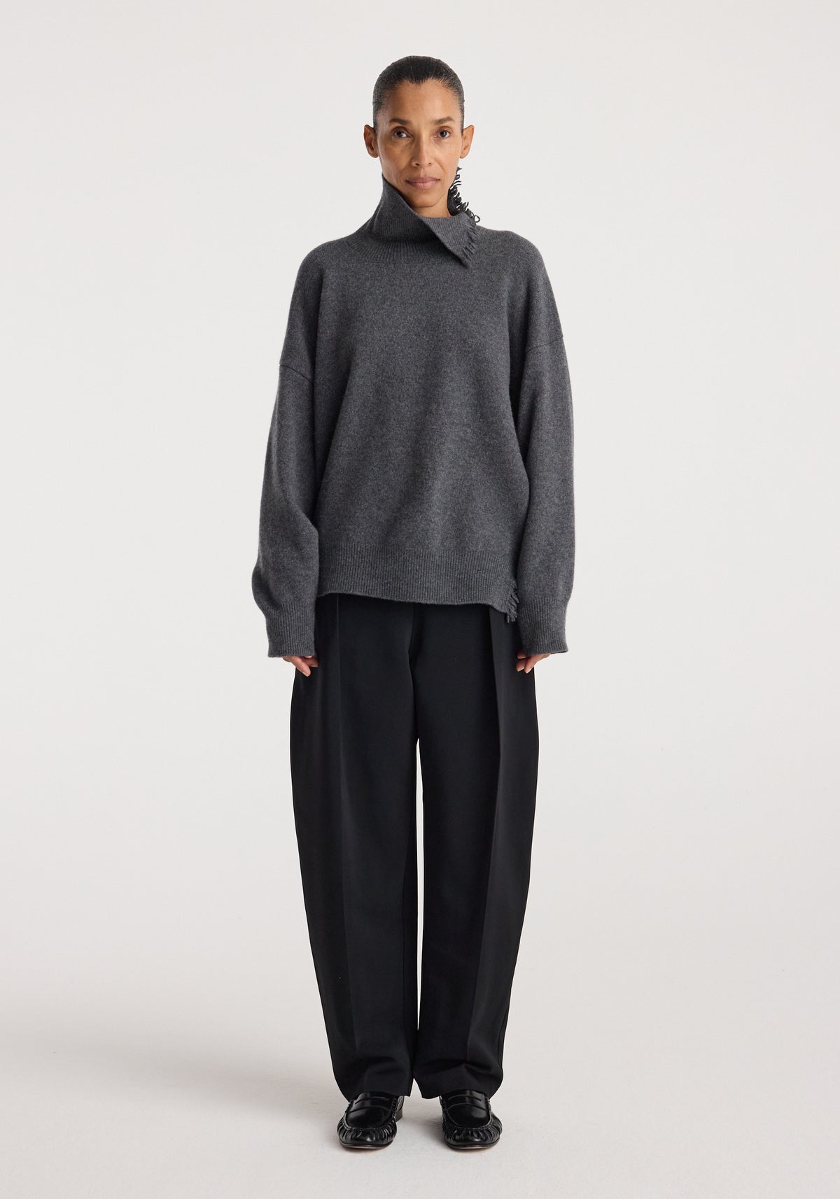 Woman wearing Turtleneck with raw details | dark grey melange, featuring an oversized fit and high collar, paired with loose black pants and black shoes against a plain background.