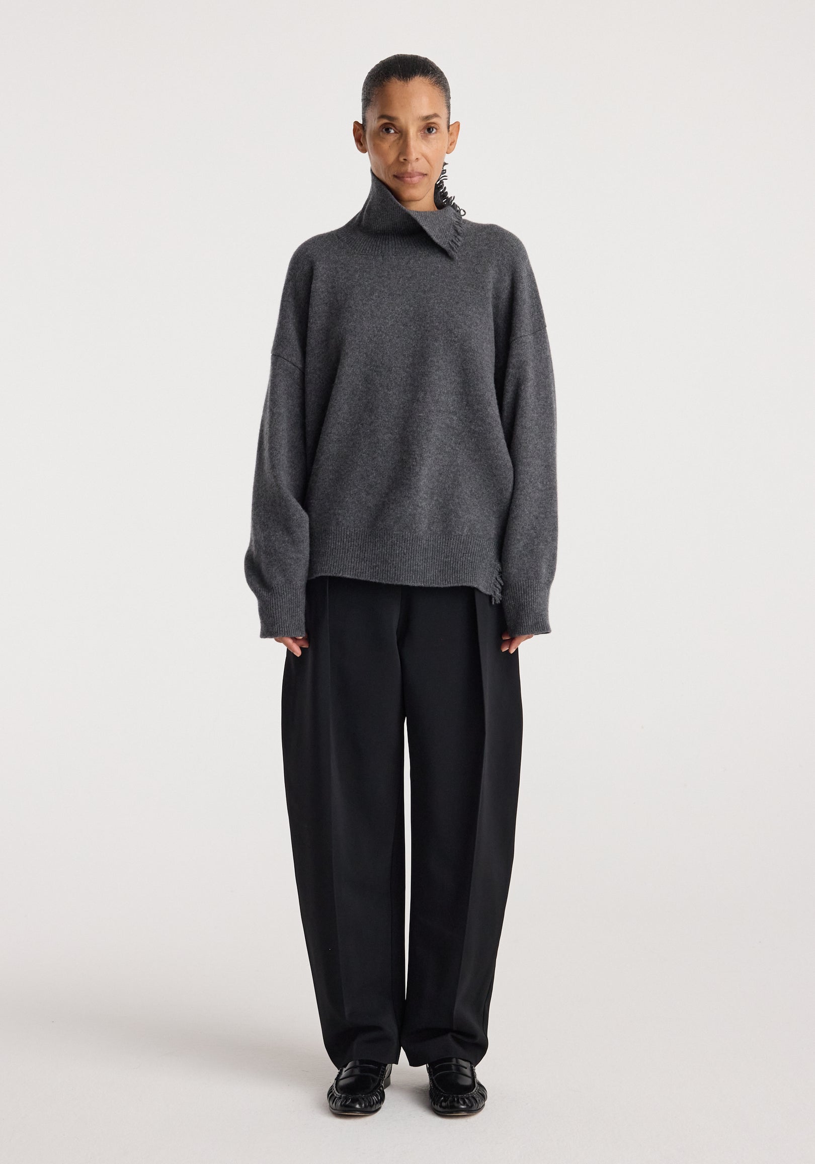 Woman wearing Turtleneck with raw details | dark grey melange, featuring an oversized fit and high collar, paired with loose black pants and black shoes against a plain background.