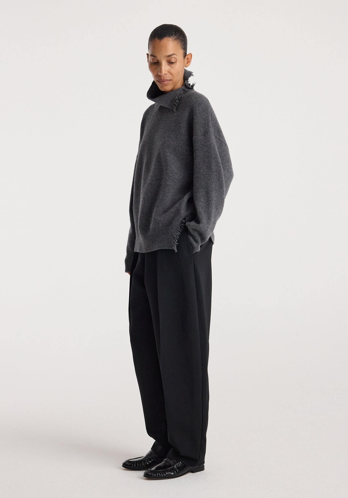 Woman wearing a dark grey melange Turtleneck with raw details, styled with loose black trousers and black shoes, posing against a plain white background.