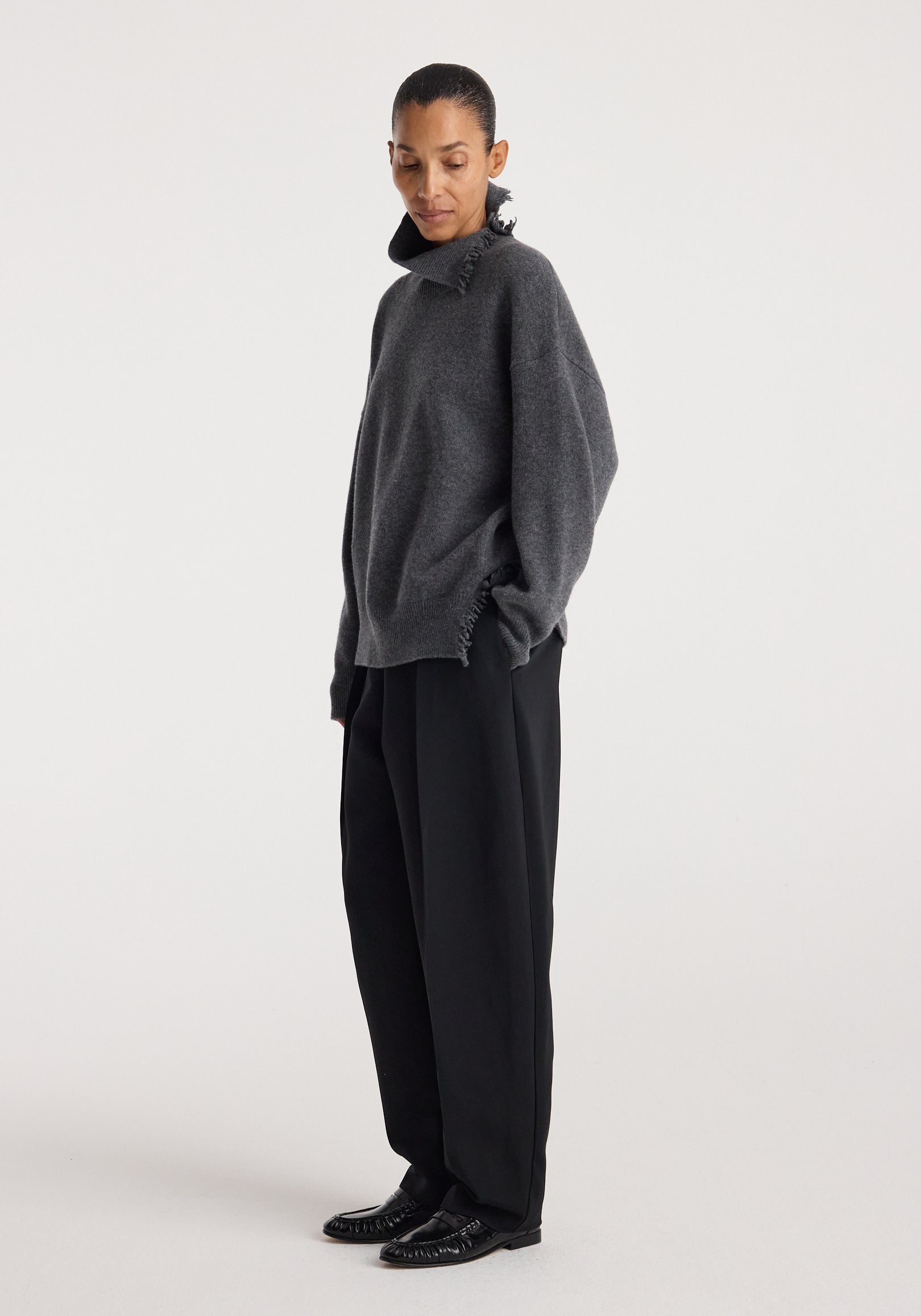 Woman wearing a dark grey melange Turtleneck with raw details, styled with loose black trousers and black shoes, posing against a plain white background.