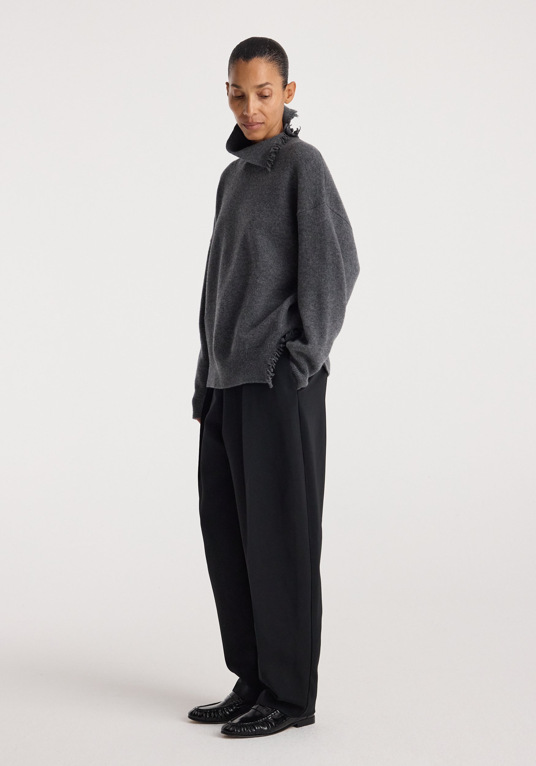 Woman wearing a dark grey melange Turtleneck with raw details, styled with loose black trousers and black shoes, posing against a plain white background.