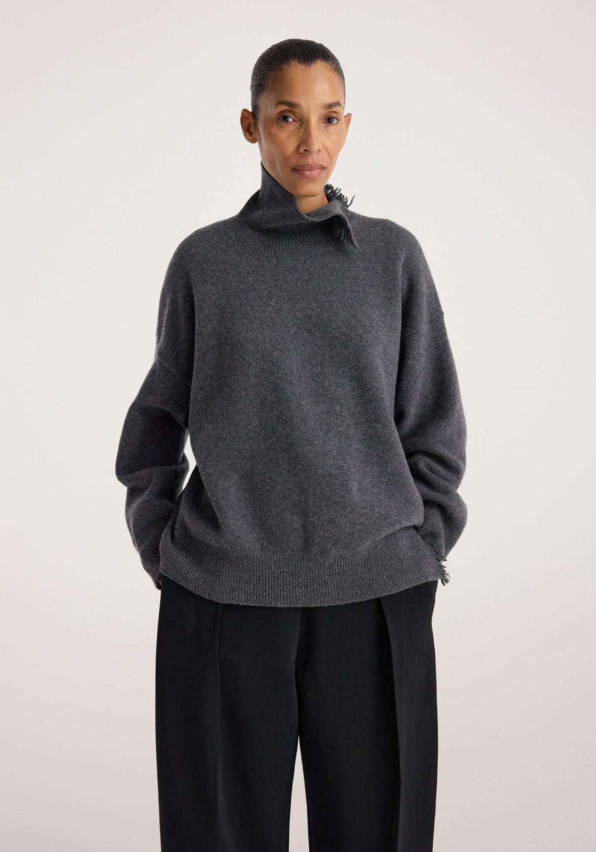 Woman wearing a dark grey melange loose-fitting Turtleneck with raw details on the collar and sleeves, paired with black wide-leg pants, standing against a plain background.