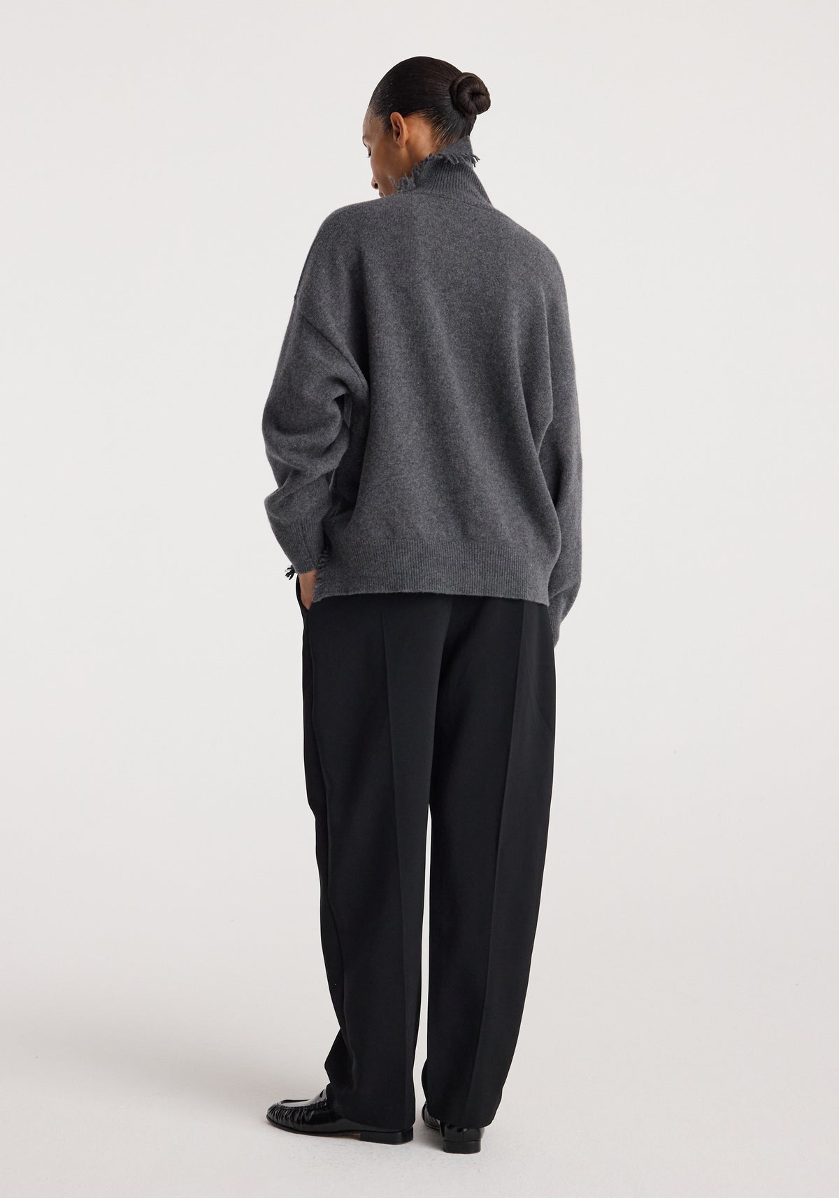 Woman wearing a Turtleneck with raw details | dark grey melange, styled with loose black pants, shown from the back against a plain background.