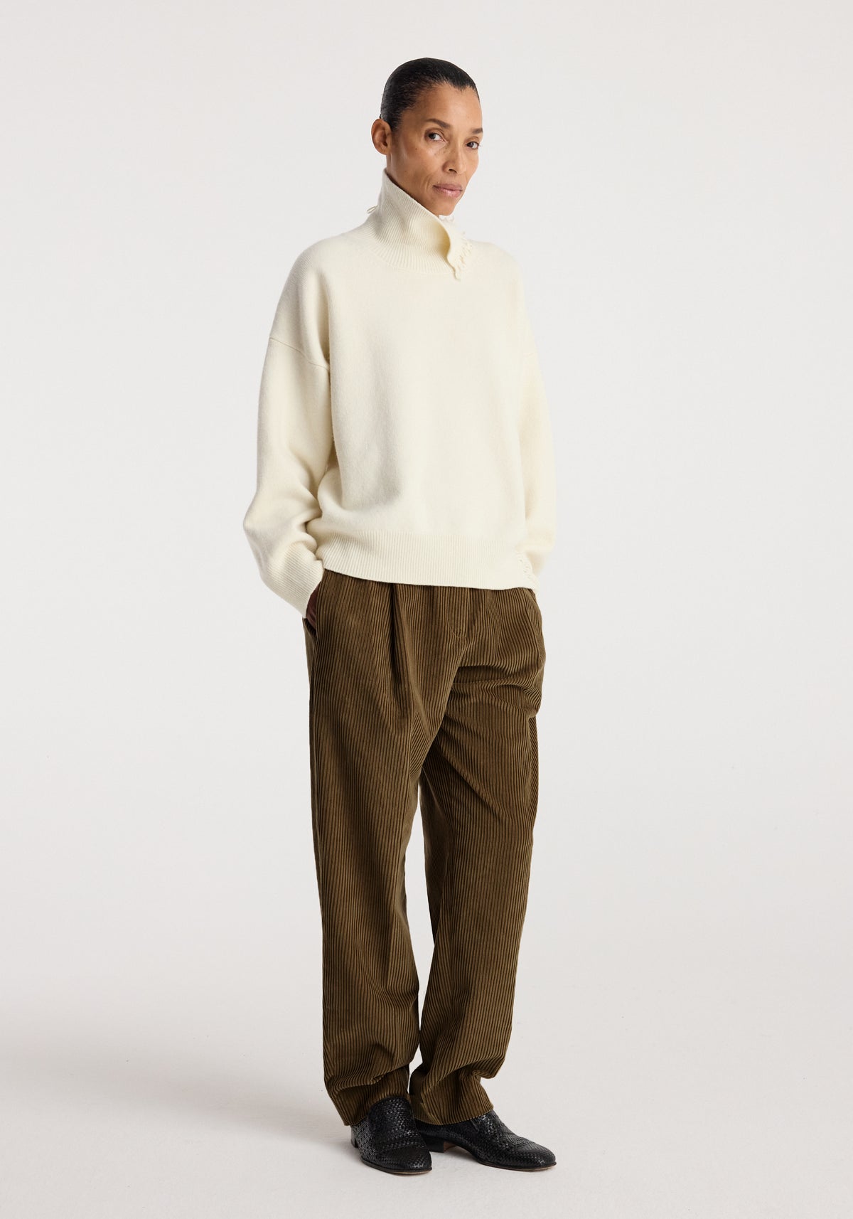 Woman wearing a Turtleneck with raw details | off white paired with loose brown corduroy pants and black shoes, standing against a plain light background.