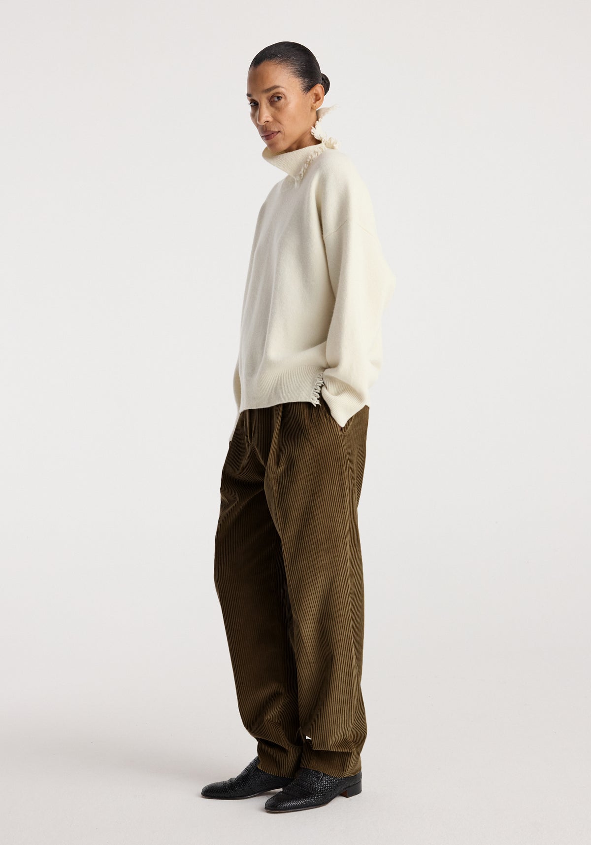 Woman wearing a Turtleneck with raw details | off white paired with loose brown corduroy pants and black shoes, standing against a plain light background.
