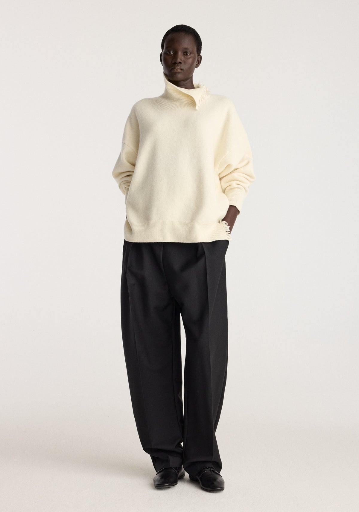 Model wearing Turtleneck with raw details | off white, a relaxed ivory sweater with rolled sleeves, paired with black trousers in a studio setting