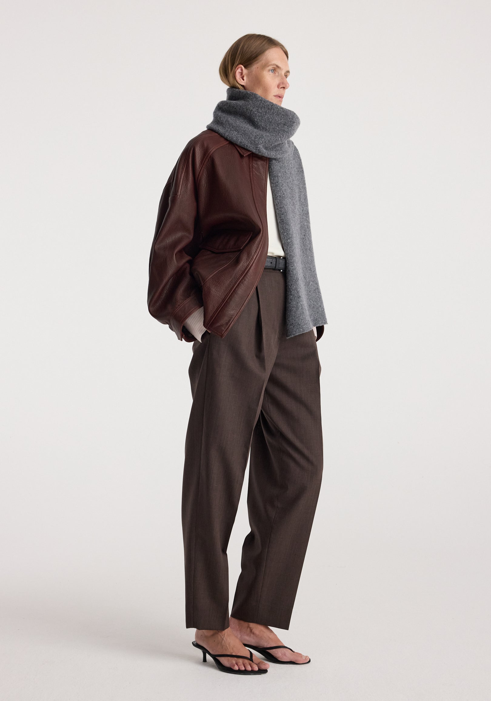 Woman wearing a gray knitted mouliné scarf | anthracite melange wrapped around her neck paired with a brown leather jacket, dark trousers, and black heeled sandals.