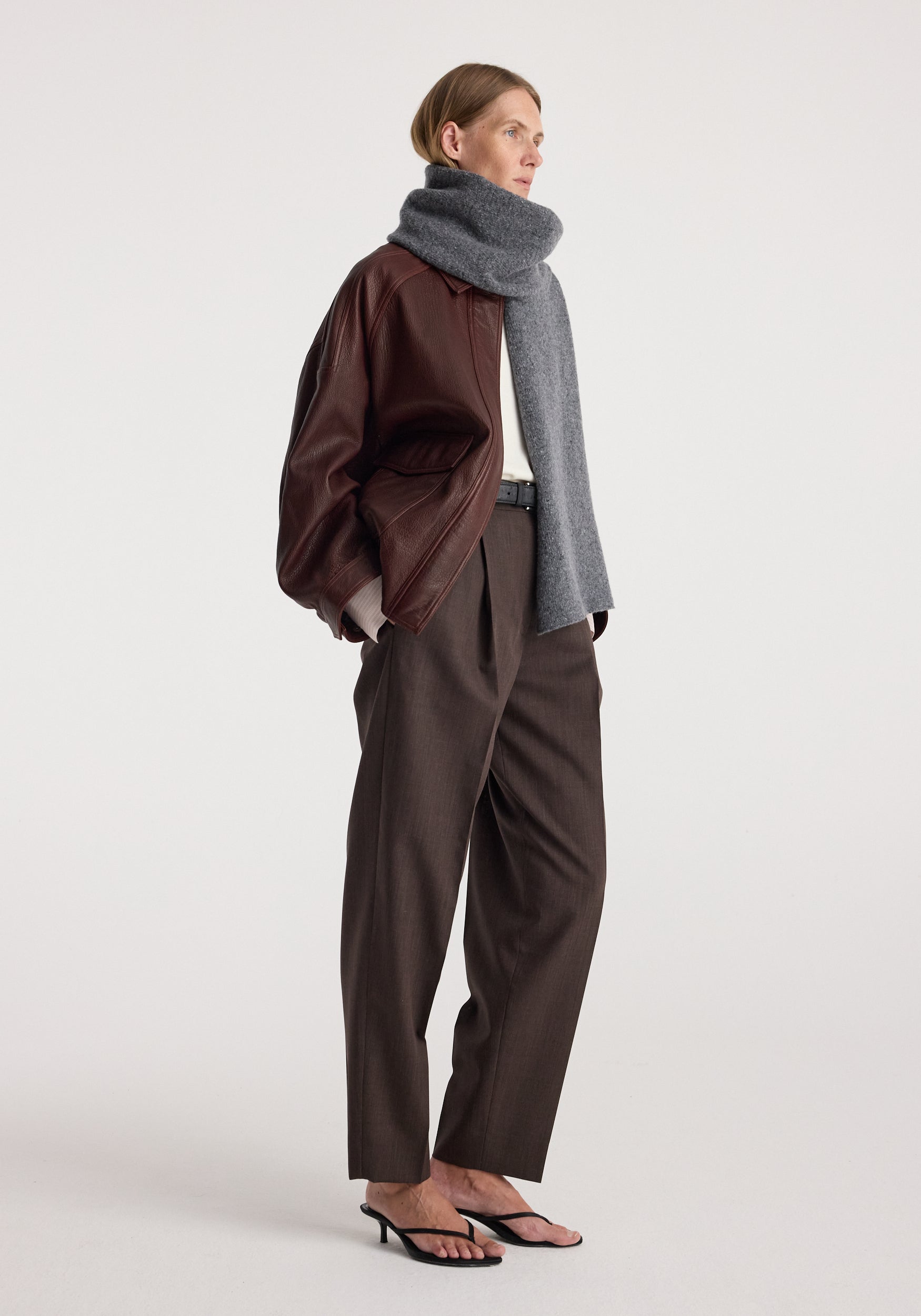 Woman wearing a gray knitted mouliné scarf | anthracite melange wrapped around her neck paired with a brown leather jacket, dark trousers, and black heeled sandals.