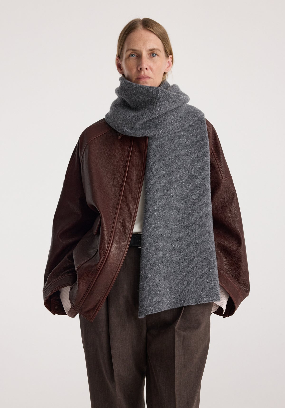 Woman wearing a thick grey knitted mouliné scarf | anthracite melange wrapped around her neck, paired with a brown leather jacket and brown pants against a plain background.