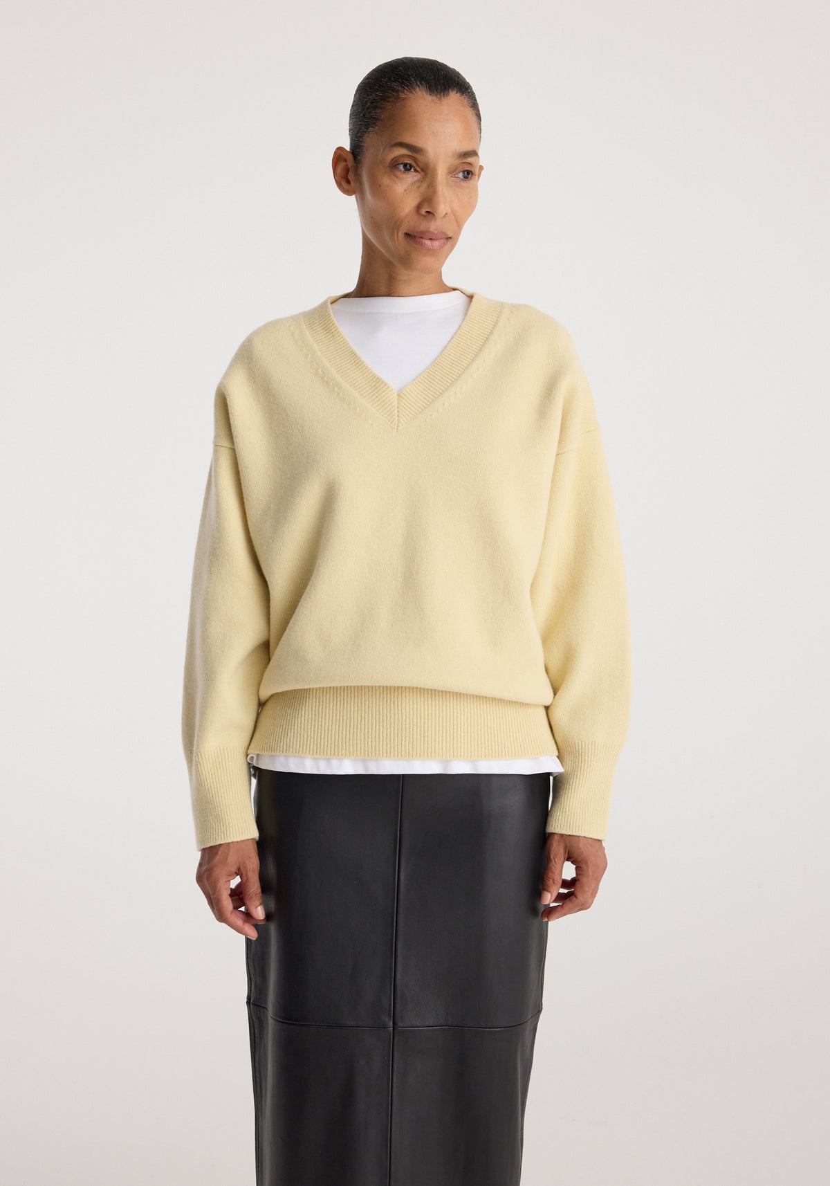 Woman wearing a Classic v-neck jumper | butter in a soft pale yellow shade with long sleeves, paired with a white shirt underneath and a black skirt against a plain background.