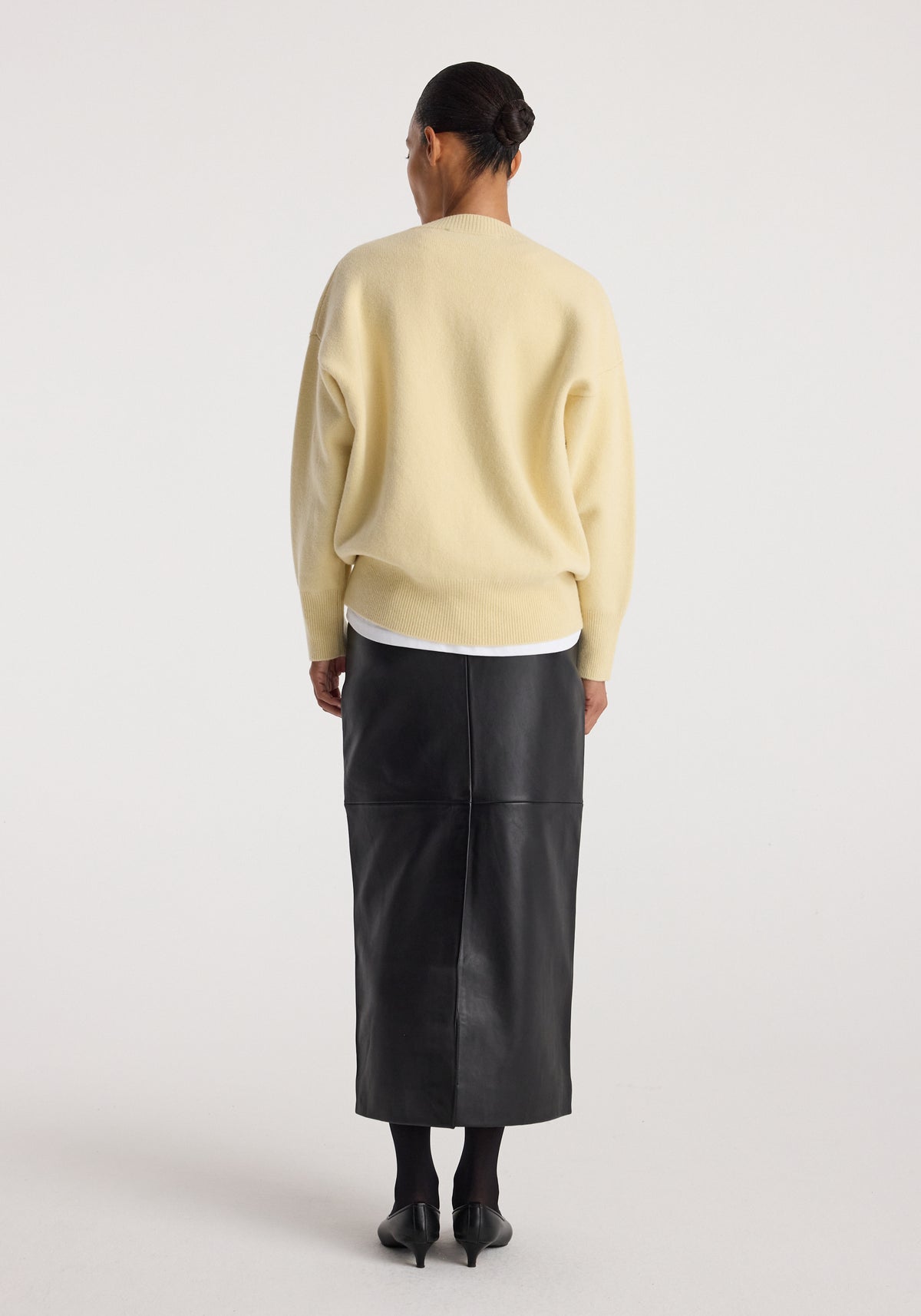Woman wearing a Classic v-neck jumper | butter in a soft yellow shade, shown from the back paired with a black midi skirt and black heels against a plain light background.
