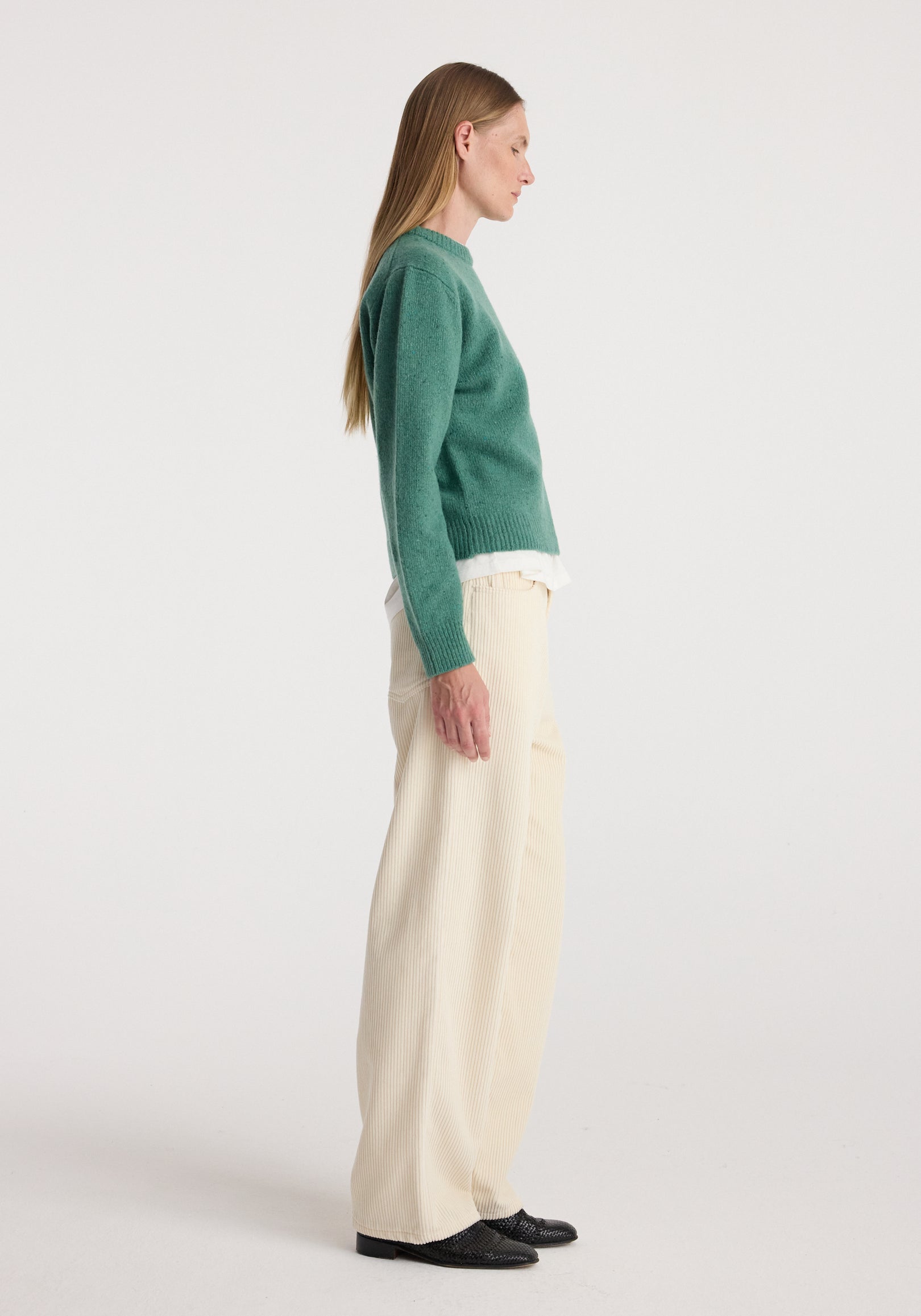 Side view of a model wearing the Fitted mouliné crewneck | jade mouliné in green with cream trousers and black shoes against a neutral background
