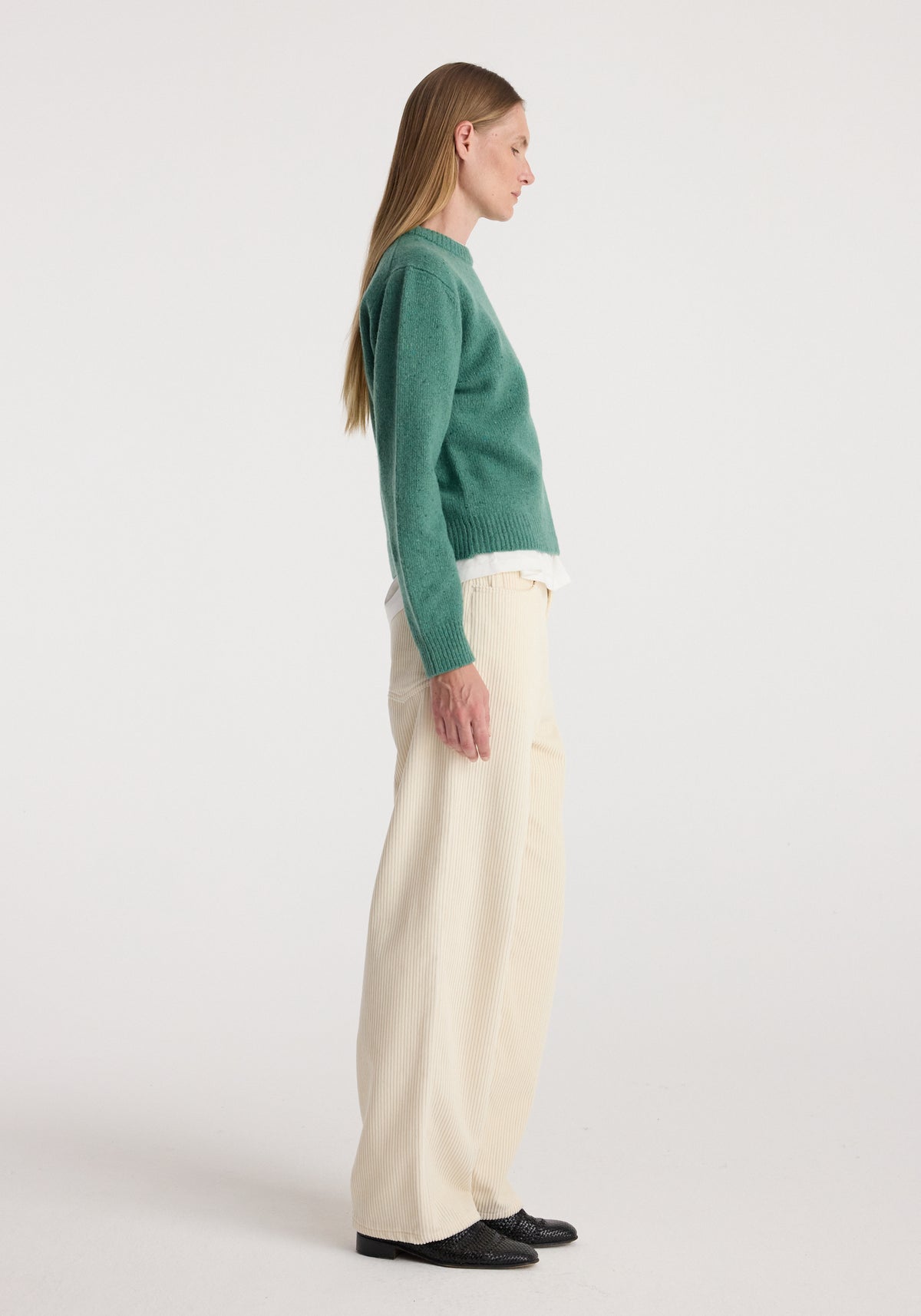 Side view of a model wearing the Fitted mouliné crewneck | jade mouliné in green with cream trousers and black shoes against a neutral background