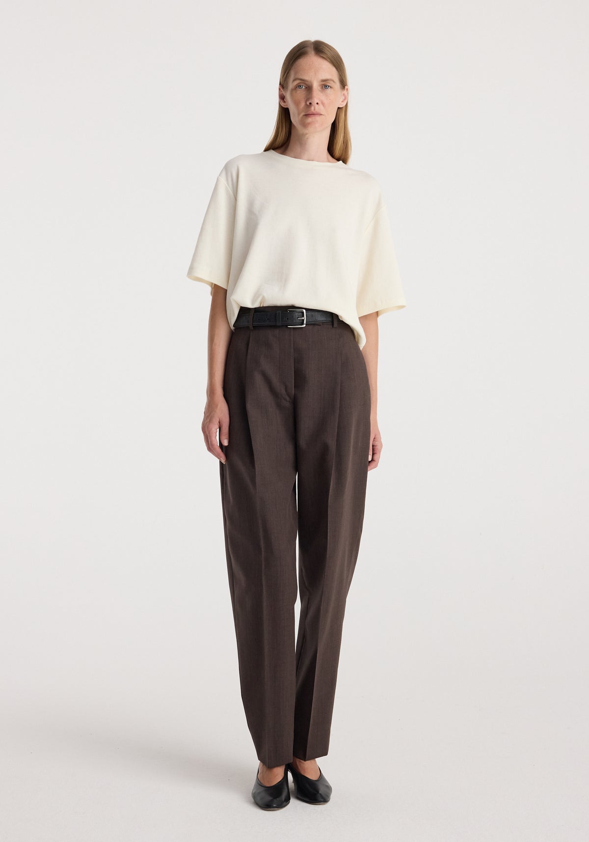 Model wearing Daria - Single pleat straight leg trousers | dark brown, styled with a cream oversized T-shirt and black shoes.