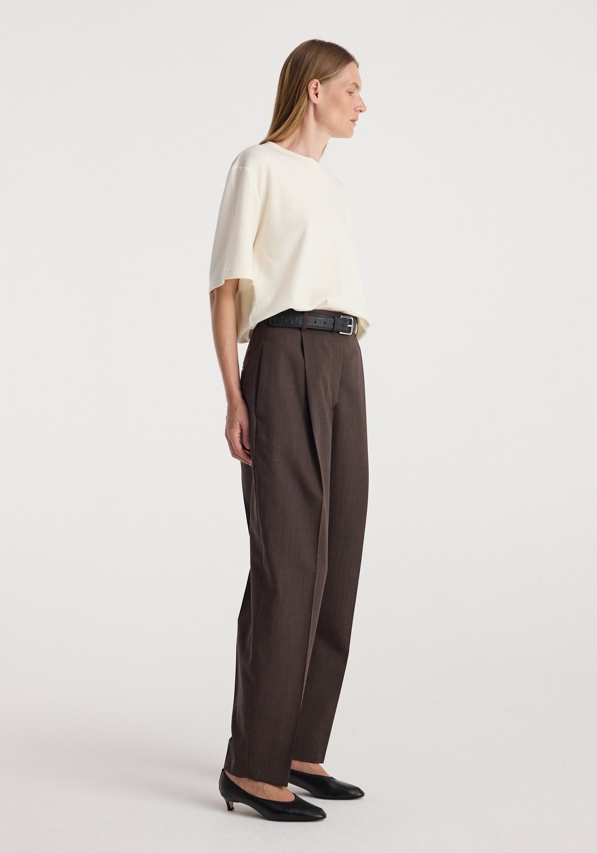 Daria - Single pleat straight leg trousers | dark brown, modeled in side profile with a cream top, black belt, and pointed black shoes.