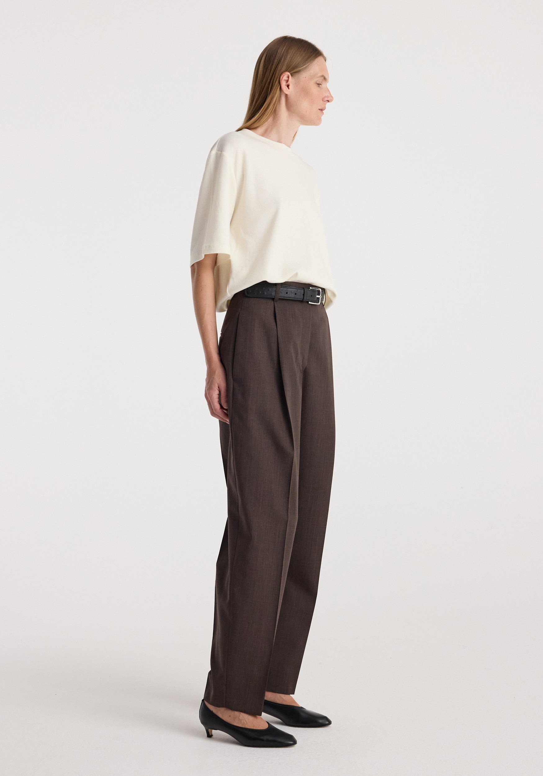 Daria - Single pleat straight leg trousers | dark brown, modeled in side profile with a cream top, black belt, and pointed black shoes.