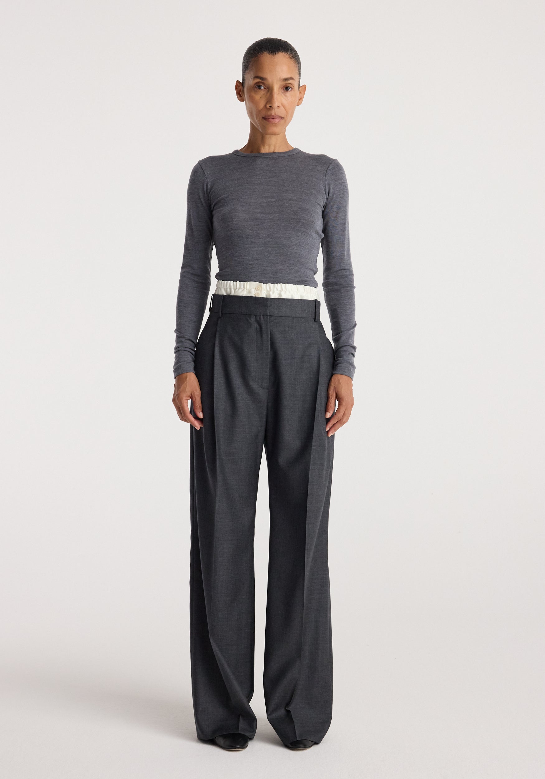 A woman wearing a fitted long-sleeve grey top paired with high-waisted, loose-fitting Boxer detailed trousers | dark grey melange, standing against a plain background.