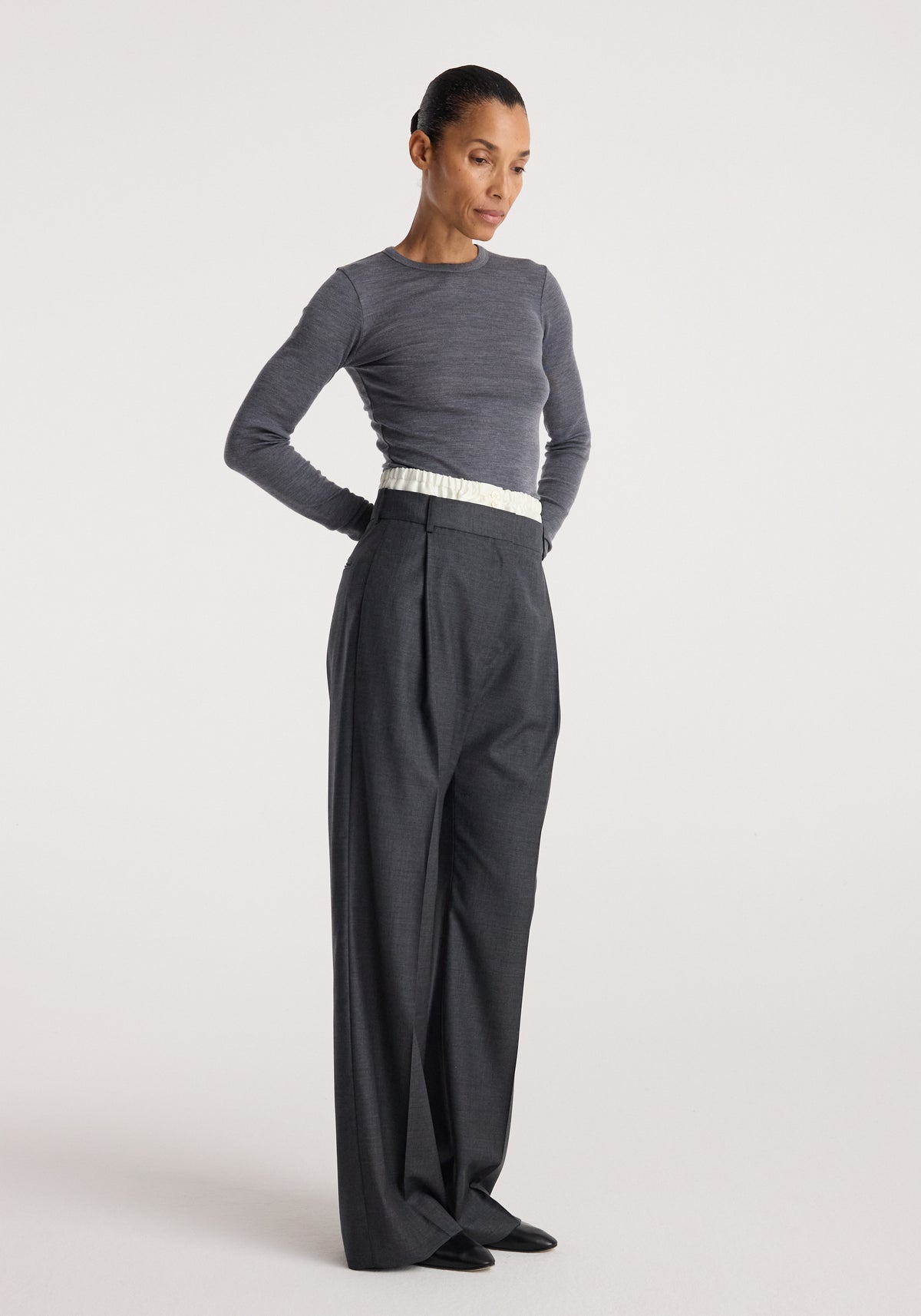 Woman wearing Boxer detailed trousers | dark grey melange with a high waist and pleated front, paired with a fitted long sleeve grey top and black shoes against a plain background.