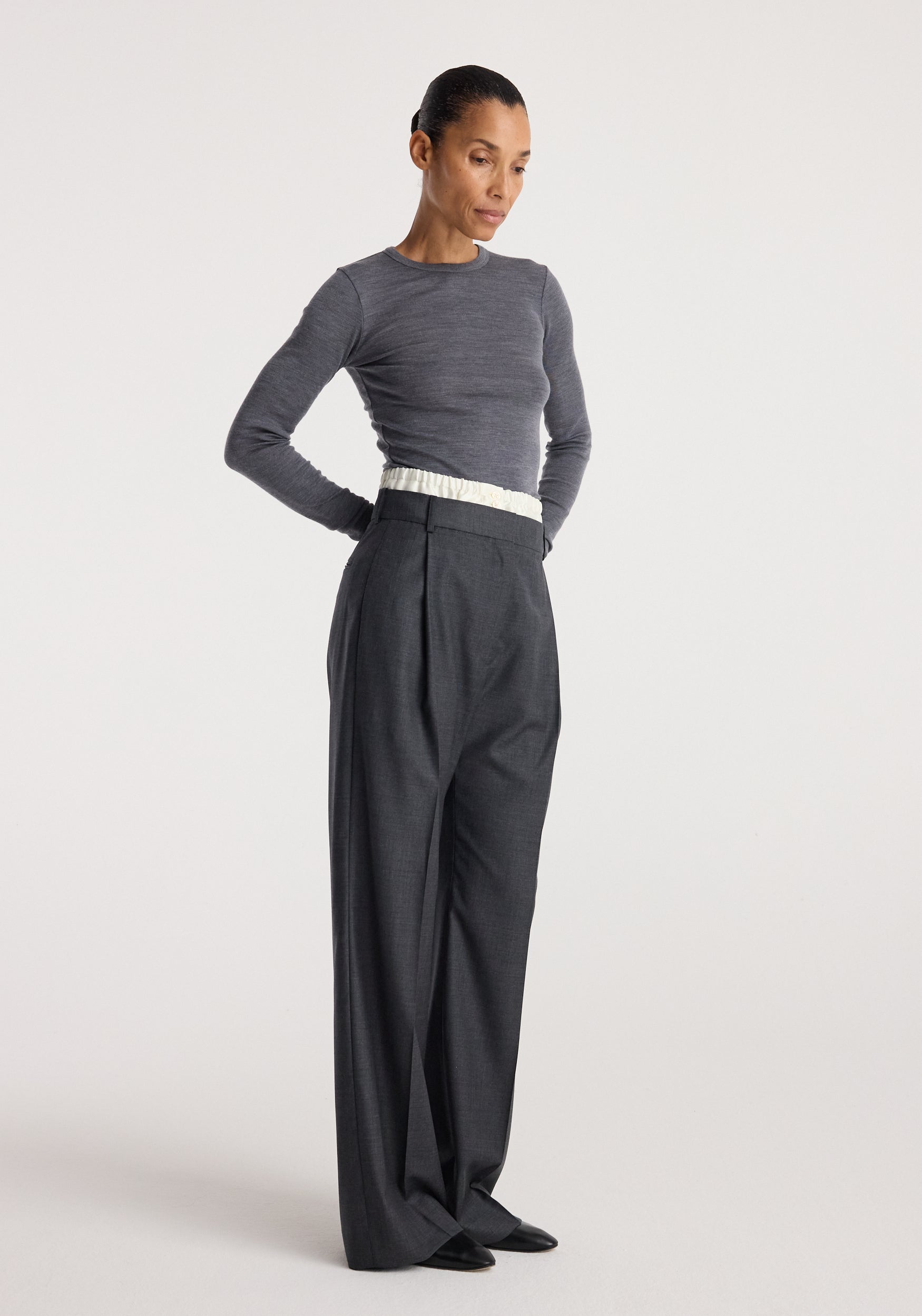 Woman wearing Boxer detailed trousers | dark grey melange with a high waist and pleated front, paired with a fitted long sleeve grey top and black shoes against a plain background.