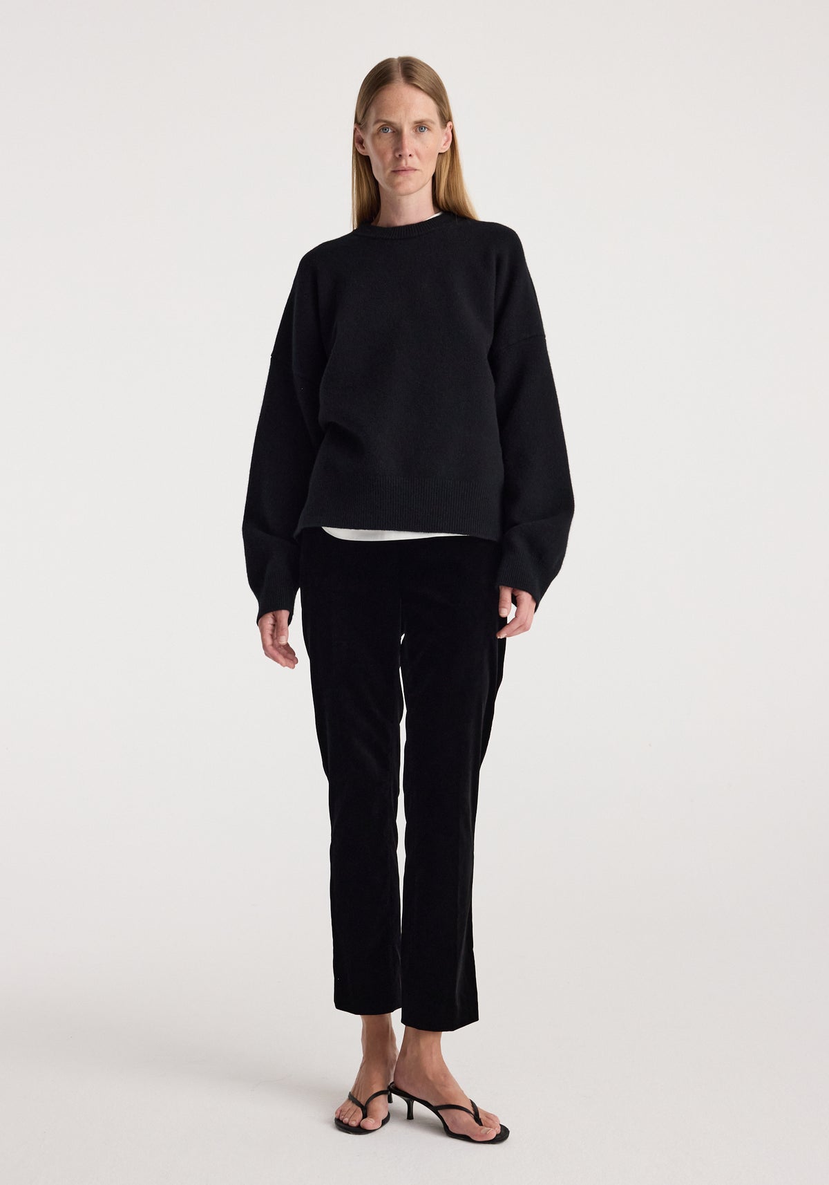 Woman wearing black Cigarette leg velvet tailored trousers | black paired with a loose-fitting black sweater and black open-toe sandals against a plain white background.