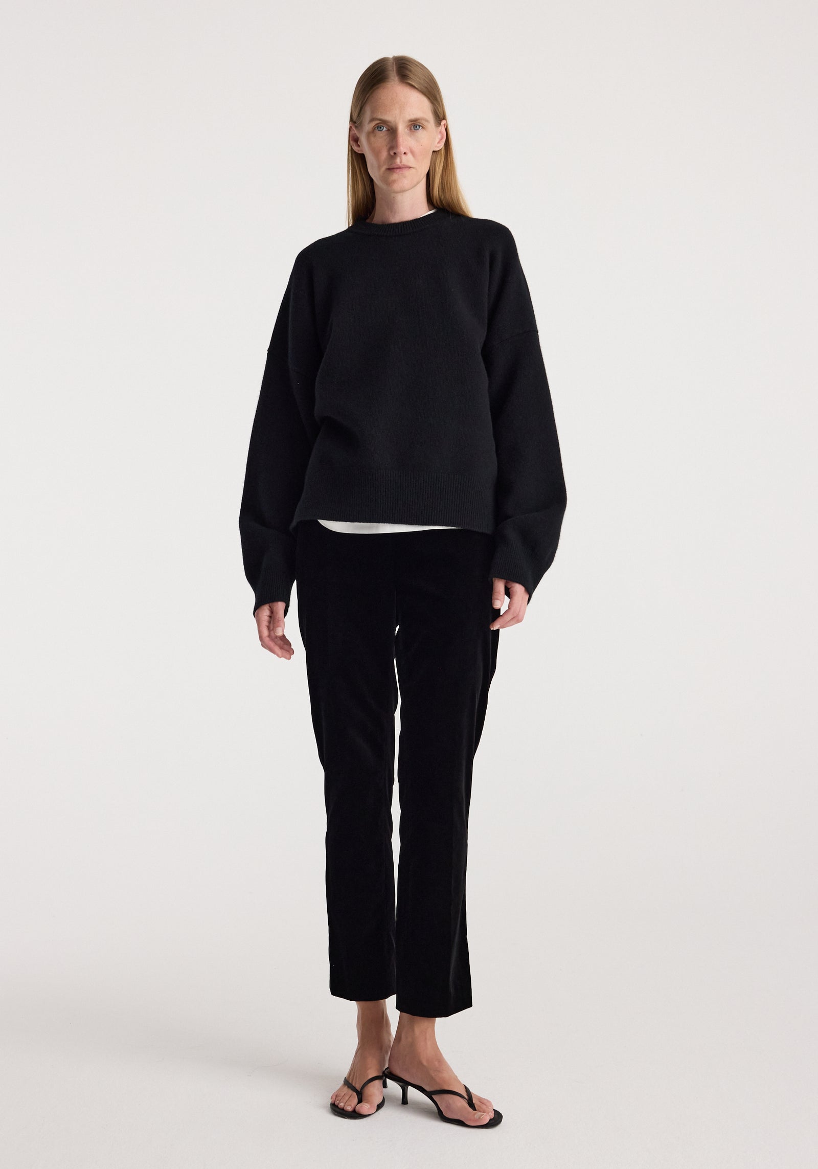 Woman wearing black Cigarette leg velvet tailored trousers | black paired with a loose-fitting black sweater and black open-toe sandals against a plain white background.
