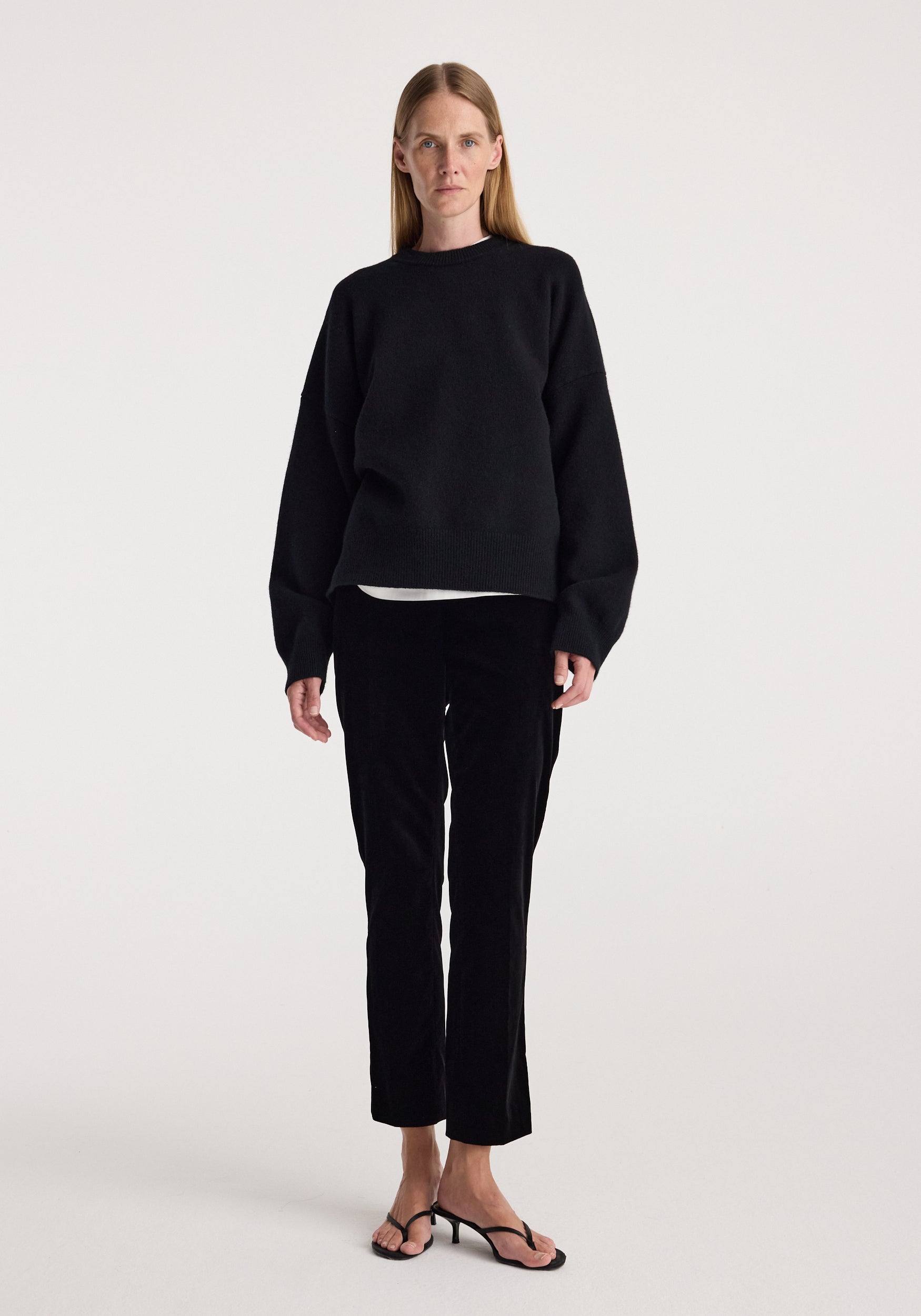Woman wearing black Cigarette leg velvet tailored trousers | black paired with a loose-fitting black sweater and black open-toe sandals against a plain white background.