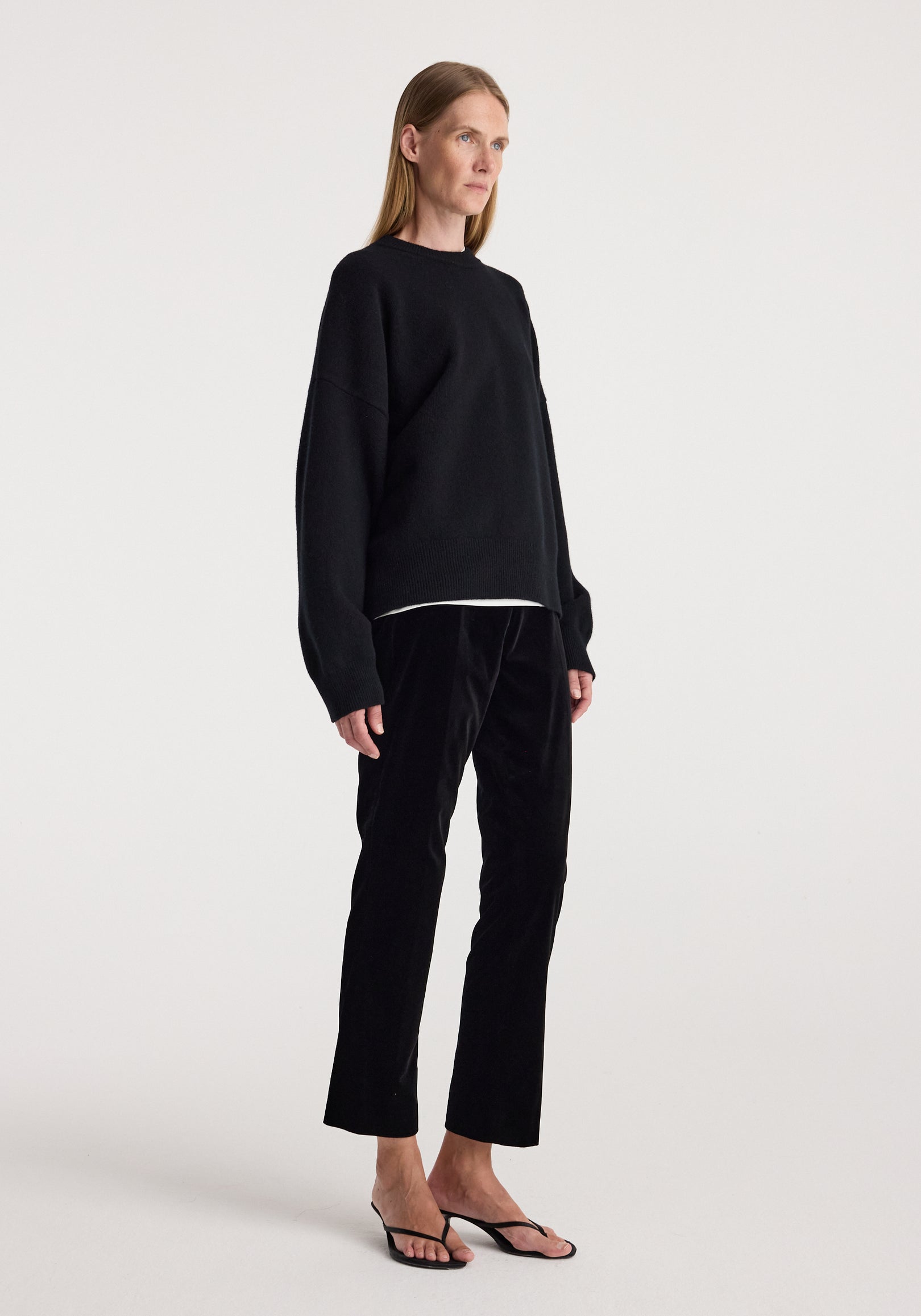 Woman wearing black Cigarette leg velvet tailored trousers | black paired with a loose black sweater and black open-toe heels against a plain background.
