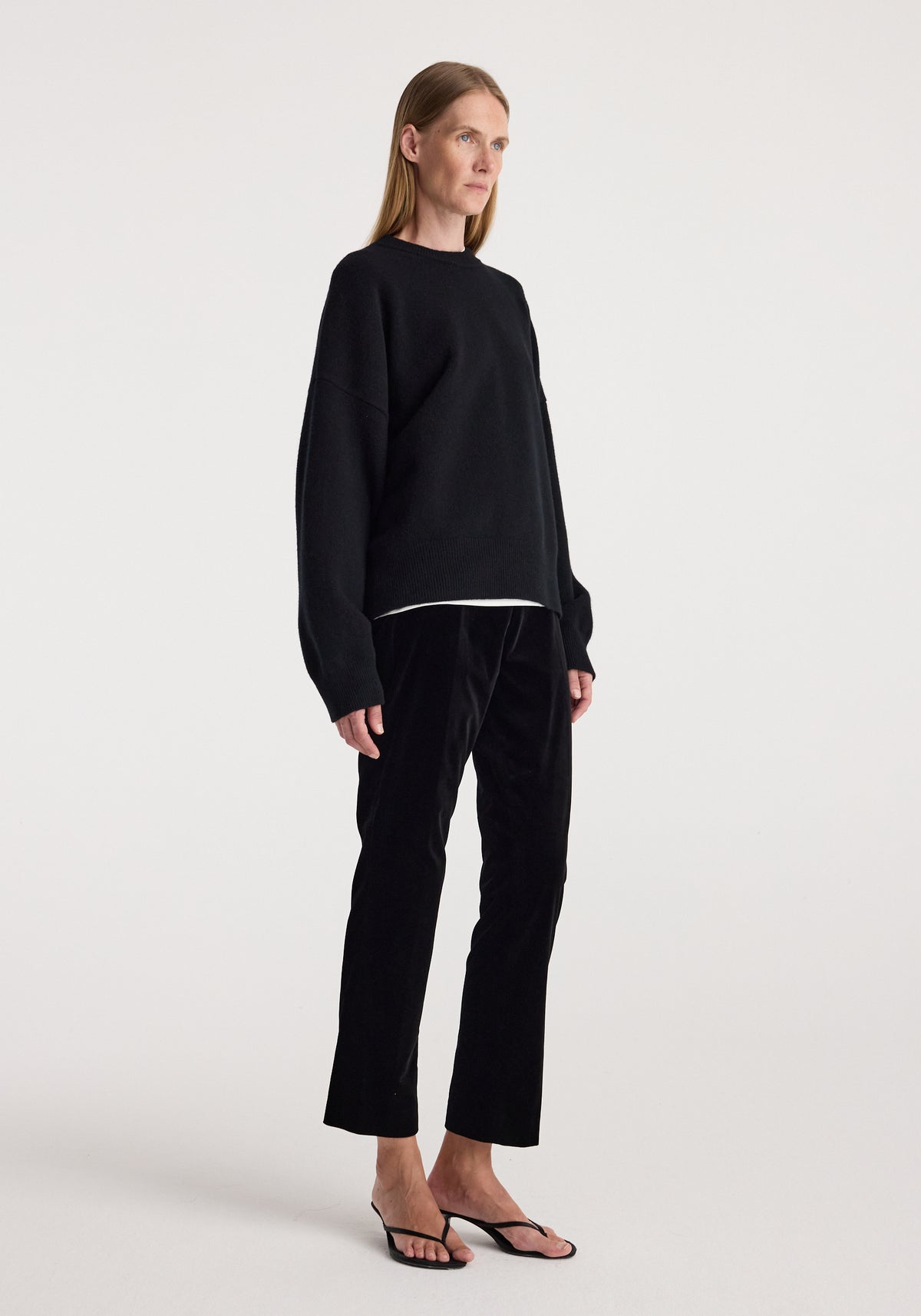 Woman wearing black Cigarette leg velvet tailored trousers | black paired with a loose black sweater and black open-toe heels against a plain background.