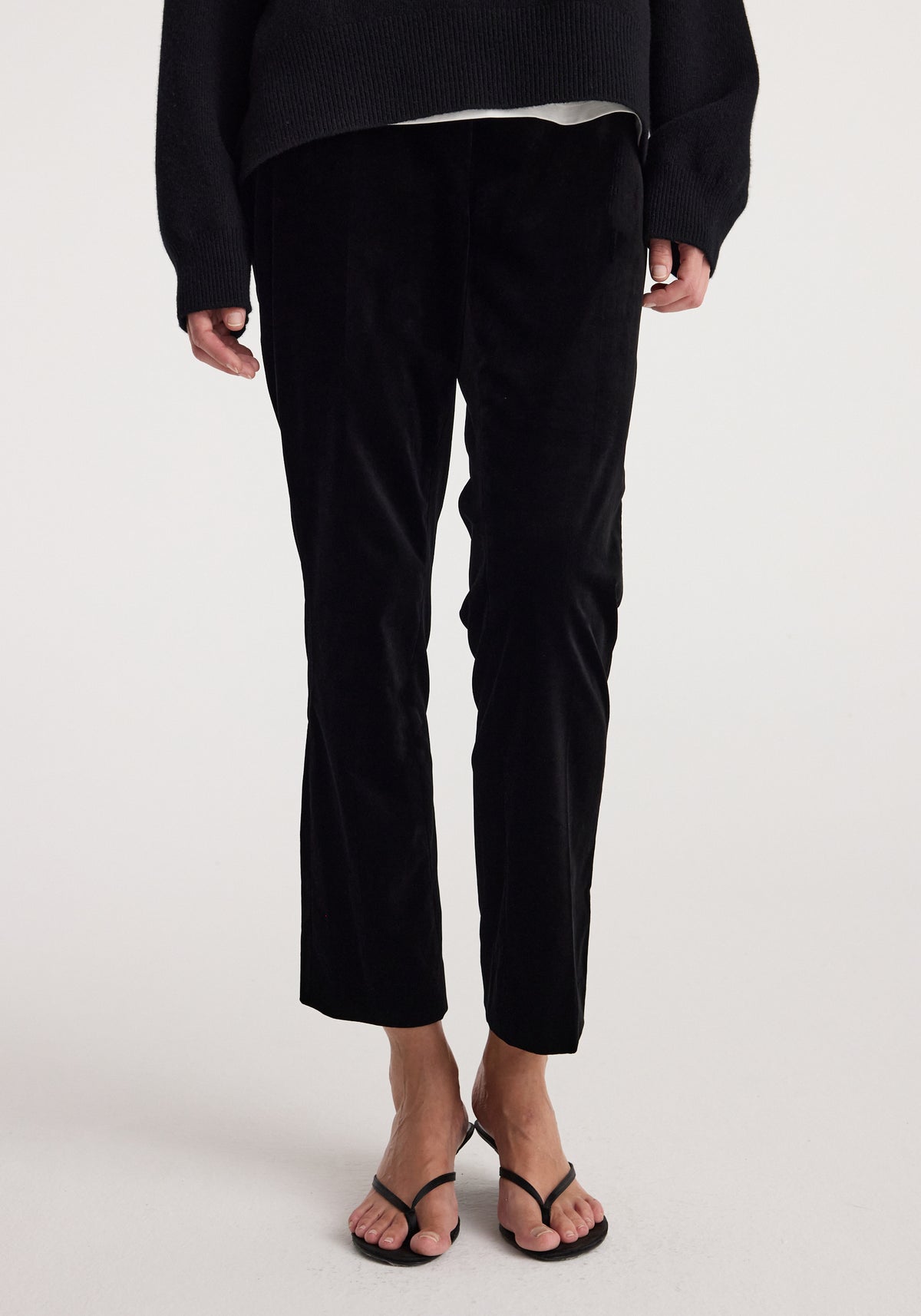 Woman wearing black Cigarette leg velvet tailored trousers | black with a fitted silhouette and a slightly cropped length paired with black strappy sandals and a dark sweater.