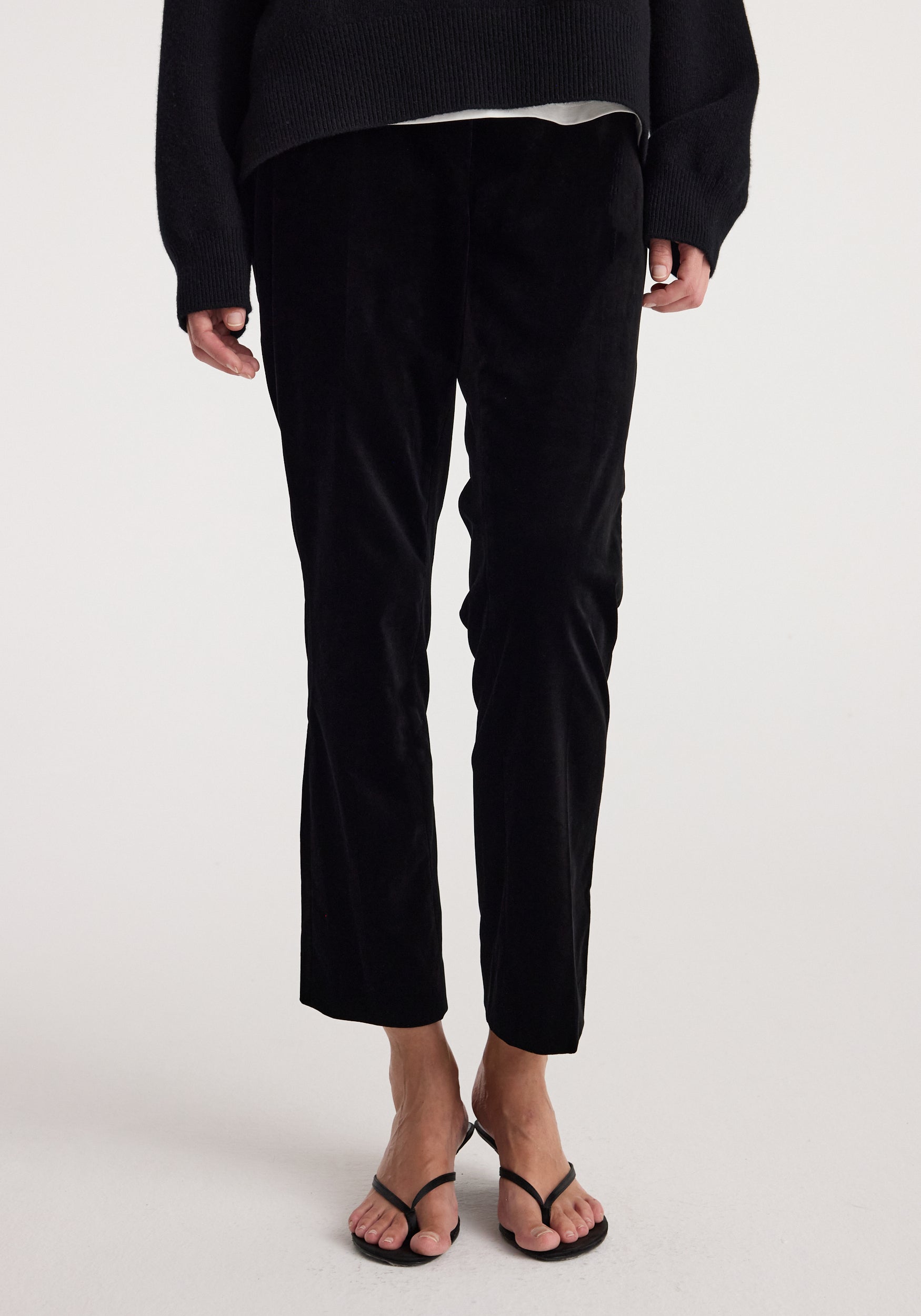 Woman wearing black Cigarette leg velvet tailored trousers | black with a fitted silhouette and a slightly cropped length paired with black strappy sandals and a dark sweater.