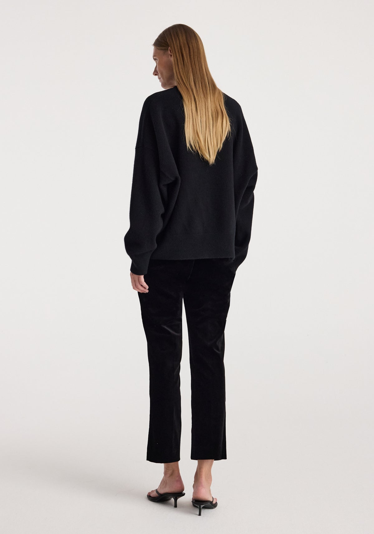 Rear view of a woman wearing black Cigarette leg velvet tailored trousers | black paired with a loose black sweater and black heeled sandals against a plain background.