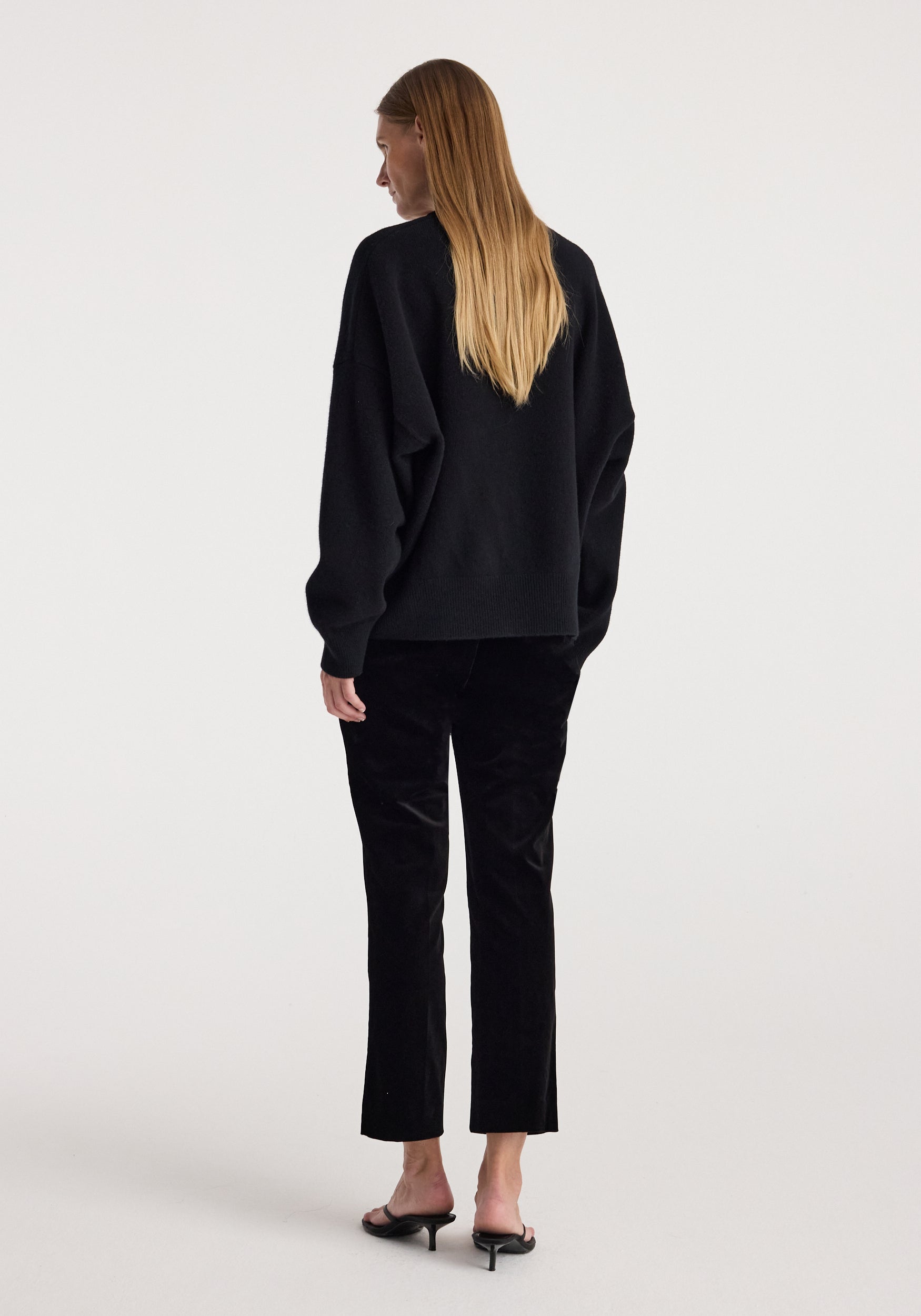 Rear view of a woman wearing black Cigarette leg velvet tailored trousers | black paired with a loose black sweater and black heeled sandals against a plain background.