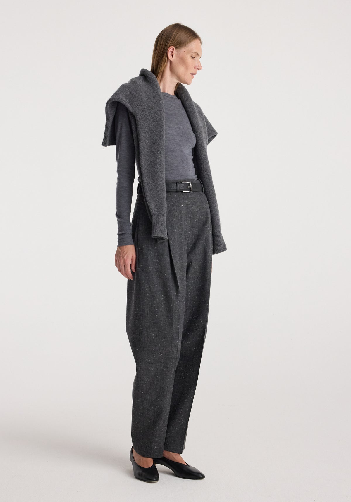A woman wearing Daria - Single pleat straight leg trousers | grey slub paired with a long-sleeve fitted grey top, a dark grey sweater draped over her shoulders, a black belt, and black flats.