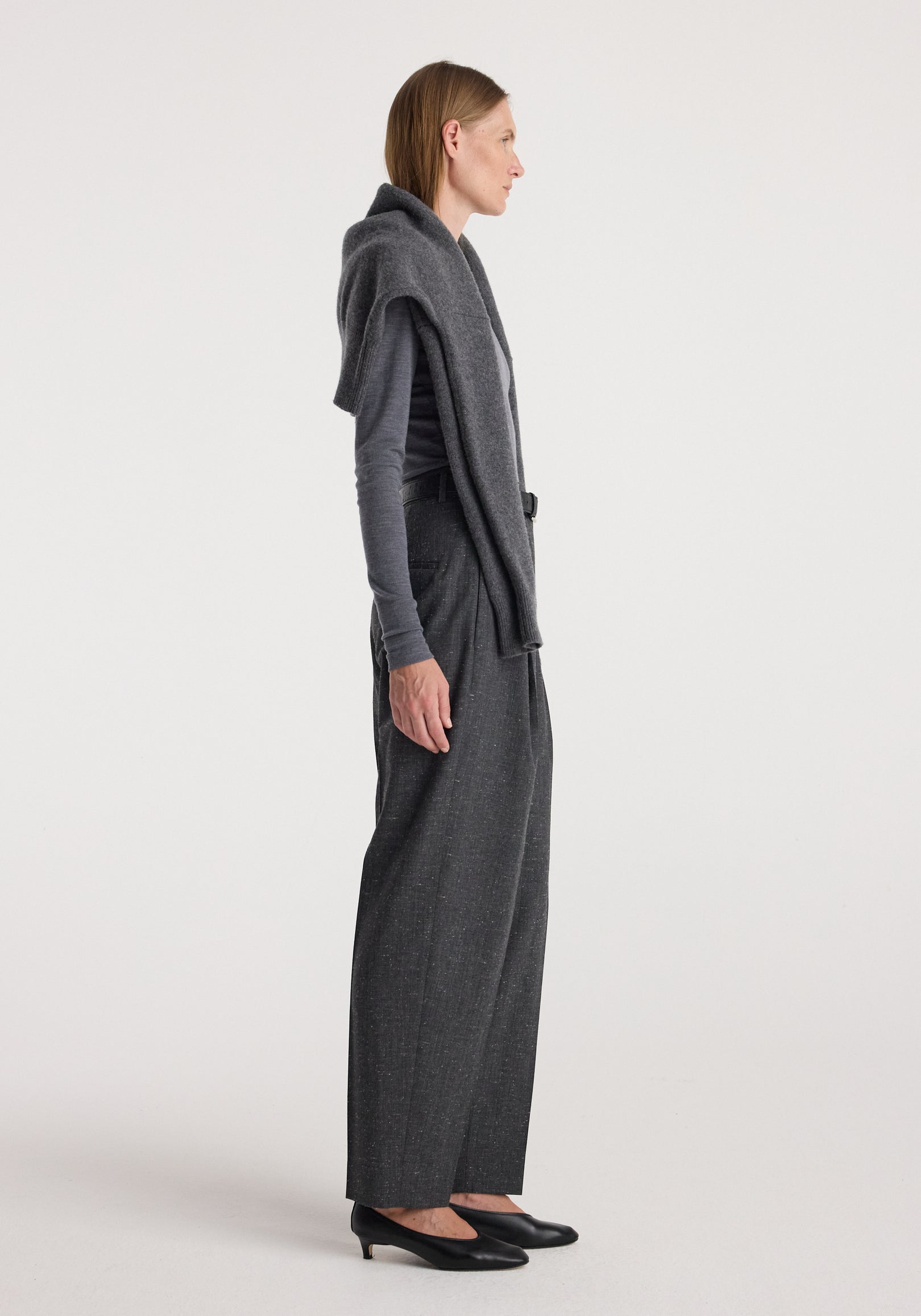 Side view of a woman wearing Daria - Single pleat straight leg trousers | grey slub with a grey long-sleeve top and a matching sweater draped over her shoulders paired with black low-heeled shoes.